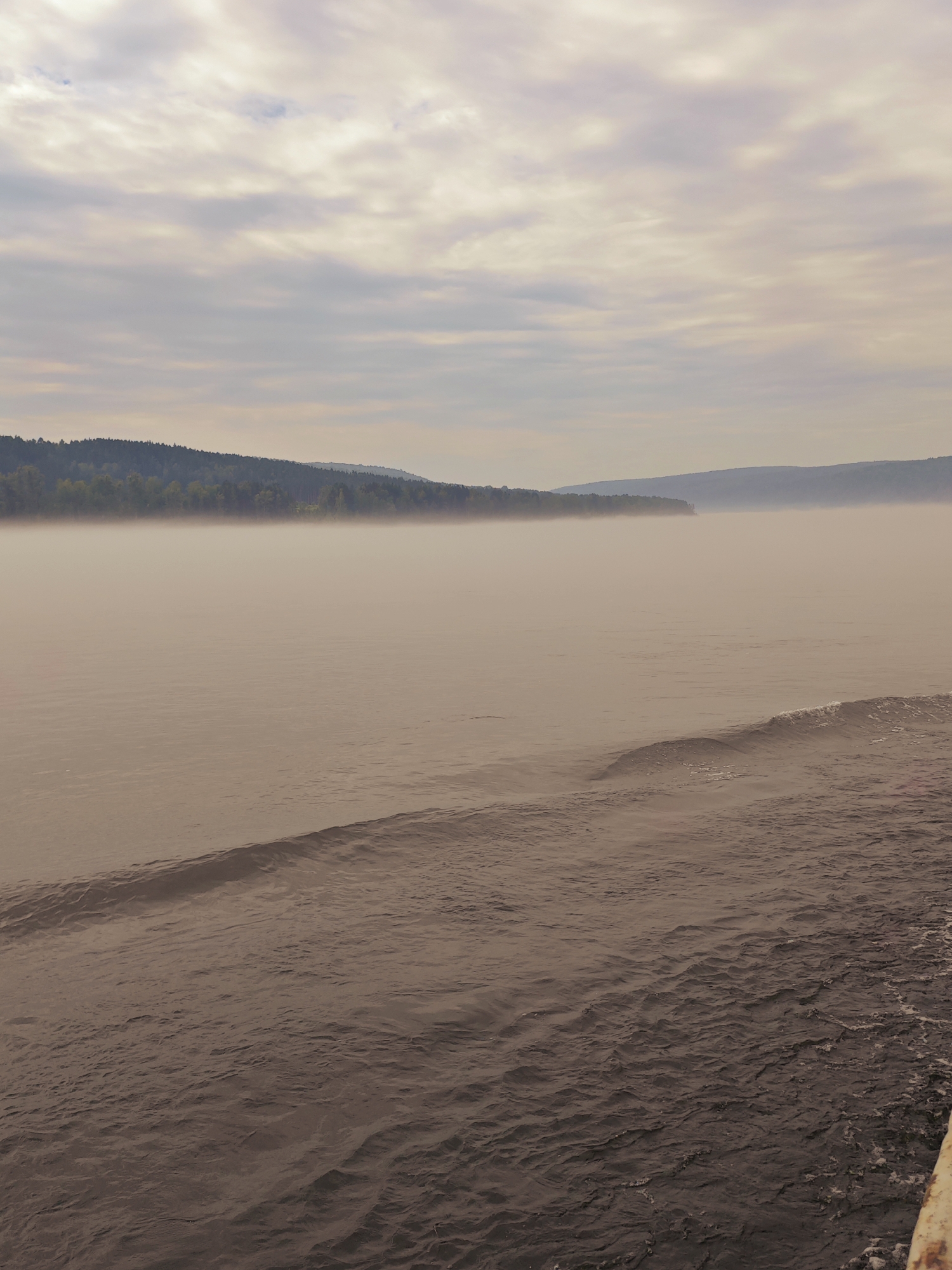 The image depicts a tranquil, misty river scene in Atamanovo, Russia. The foreground is dominated by the rippling surface of the water, showing subtle waves and a wake. In the midground, a thick layer of fog partially obscures the distant shoreline, which is lined with a dense, dark green forest. The background features rolling hills fading into the hazy distance under a cloudy sky. The overall atmosphere is serene and slightly melancholic, with muted colors and soft lighting contributing to the impression of an early morning or late evening. There are no people or man-made structures visible in the scene, emphasizing the natural beauty of the location.