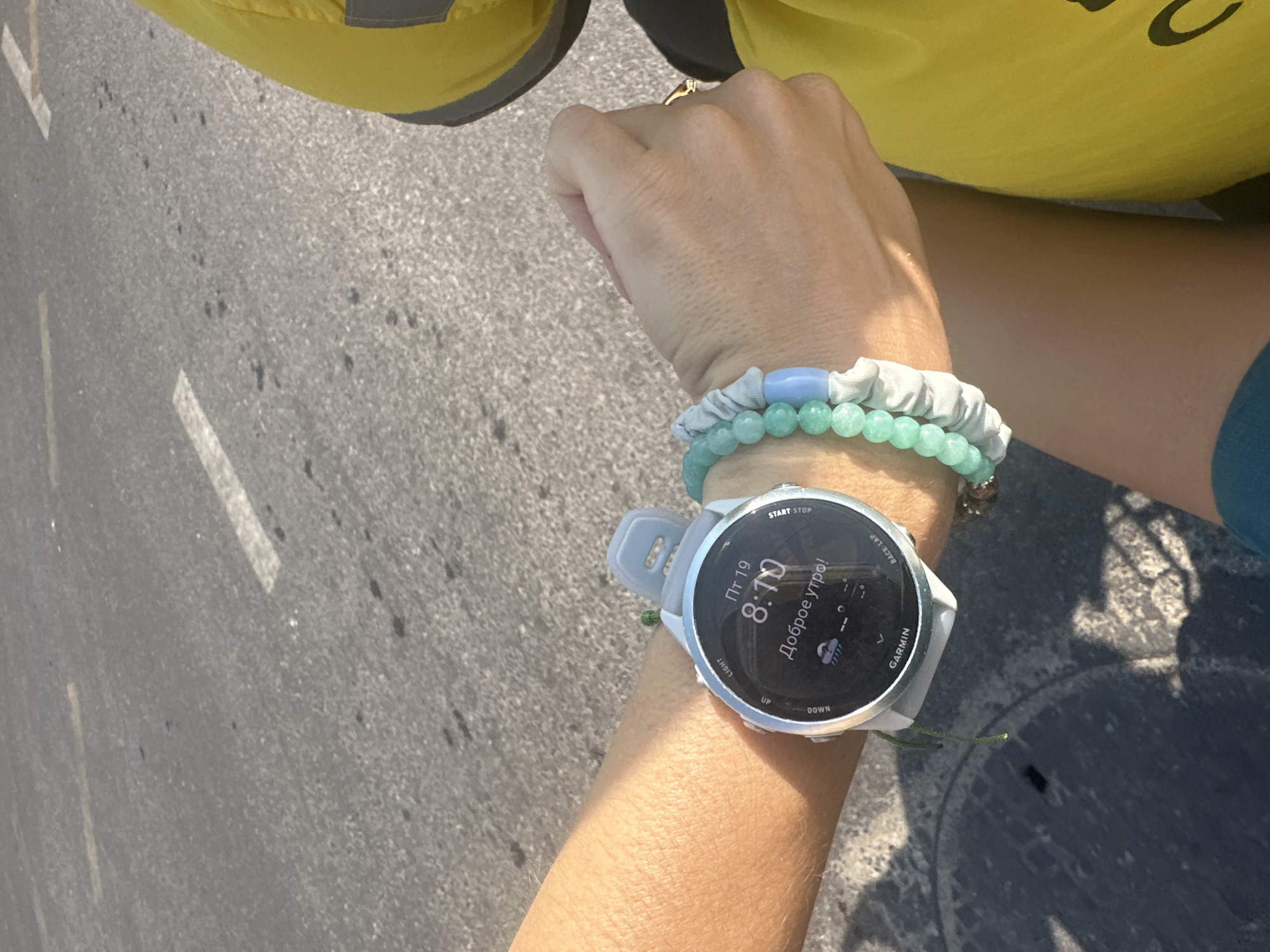 The image shows a close-up of a person's wrist wearing a Garmin watch and two beaded bracelets. The watch face displays the time as 8:10 and shows the text "Доброе утро!" which translates to "Good morning!" in Russian. It also indicates "Пт 19," likely representing Friday the 19th. A small icon suggests the weather is cloudy with some rain. The watch is light gray with a white band. The bracelets are light blue and green. The background is a paved surface, possibly a road or sidewalk, with some white markings. The person is wearing a yellow garment. The lighting suggests it is daytime. The overall impression is a snapshot of someone checking the time and greeting the day, possibly before or during an outdoor activity.