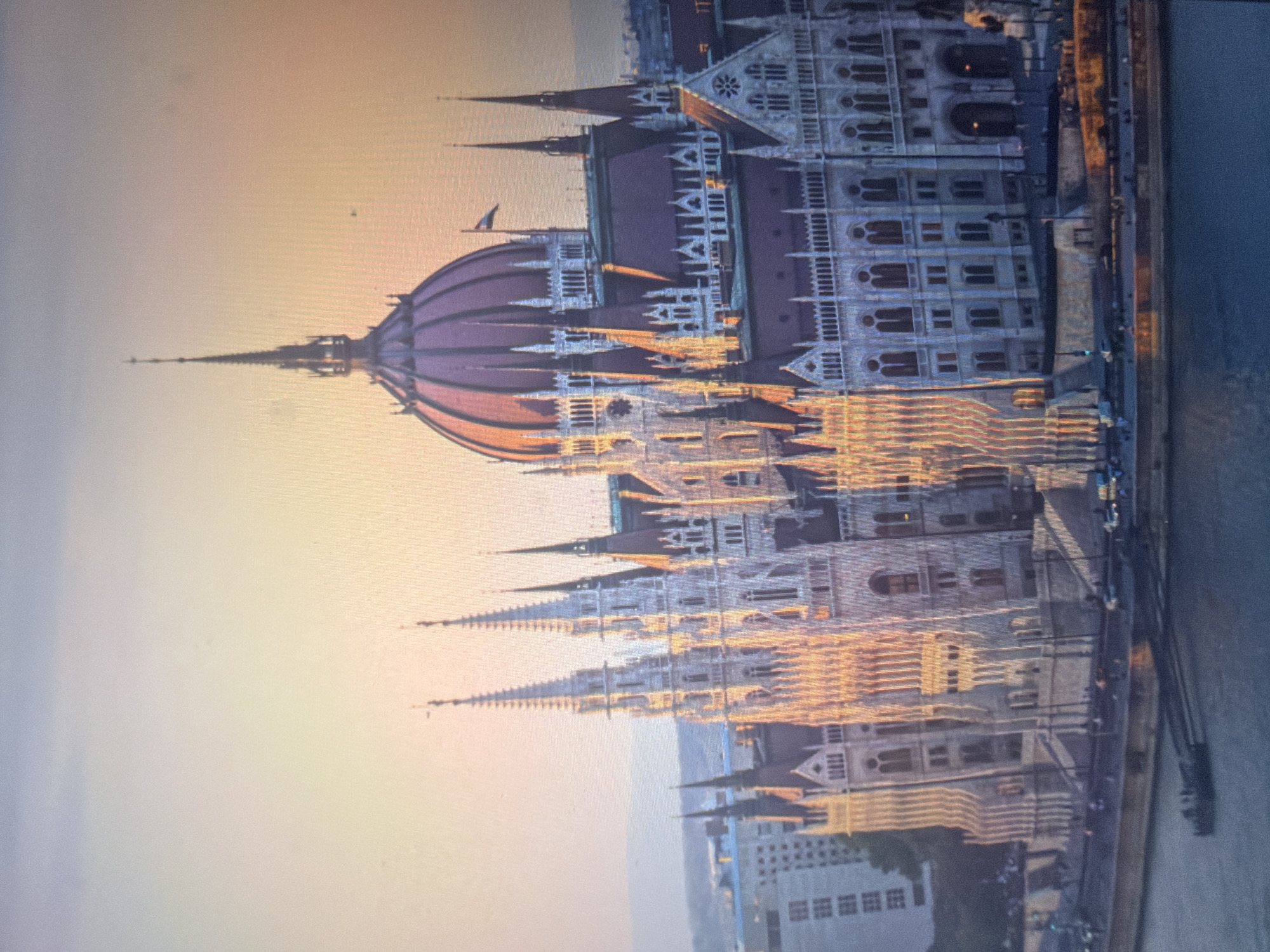 The image depicts an ornate, monumental Gothic Revival-style building, characterized by numerous pointed spires, intricate stone detailing, and a prominent central dome with a reddish roof. The structure is illuminated by warm, golden sunlight from the right, suggesting either sunrise or sunset, which casts soft highlights and shadows across its complex facade. The sky transitions from a pale blue at the top to vibrant yellow, orange, and pink hues near the horizon.
The building is situated along the edge of a wide body of water, likely a river, with a paved promenade or embankment running along its base. Indistinct figures of people and several parked vehicles are visible on this waterfront area. In the lower left foreground on the water, a dark, rectangular boat or dock structure is present. The water itself is a dark blue-grey. To the left of the main building, other, more modern city buildings are partially visible in the background, and a faint outline of distant hills or mountains can be seen on the horizon. A tricolor flag flies from one of the smaller spires near the central dome. A small dark speck, possibly a bird, is visible in the sky above the dome. This scene is located in the city of Shumen, Bulgaria.