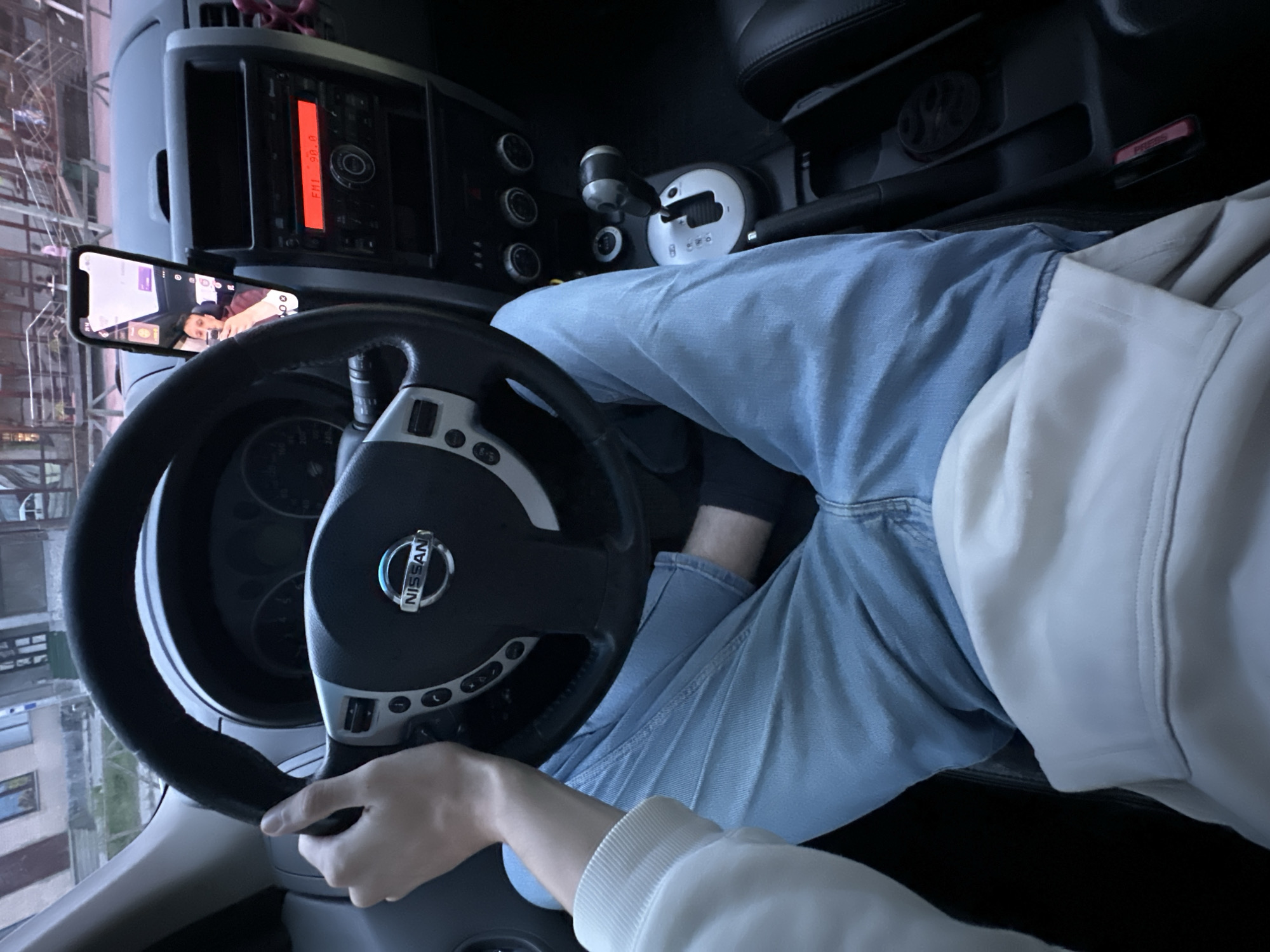 The image shows the interior of a Nissan vehicle, likely in Shatava, Ukraine. A person is seated in the driver's seat, holding the steering wheel with their left hand. They are wearing a white hoodie and light blue jeans. A smartphone is mounted on the dashboard, displaying a video call or recording of the person. The car's radio is on, showing "FM1 98.0". The surroundings visible through the windshield suggest an urban or suburban environment with buildings and streetlights.