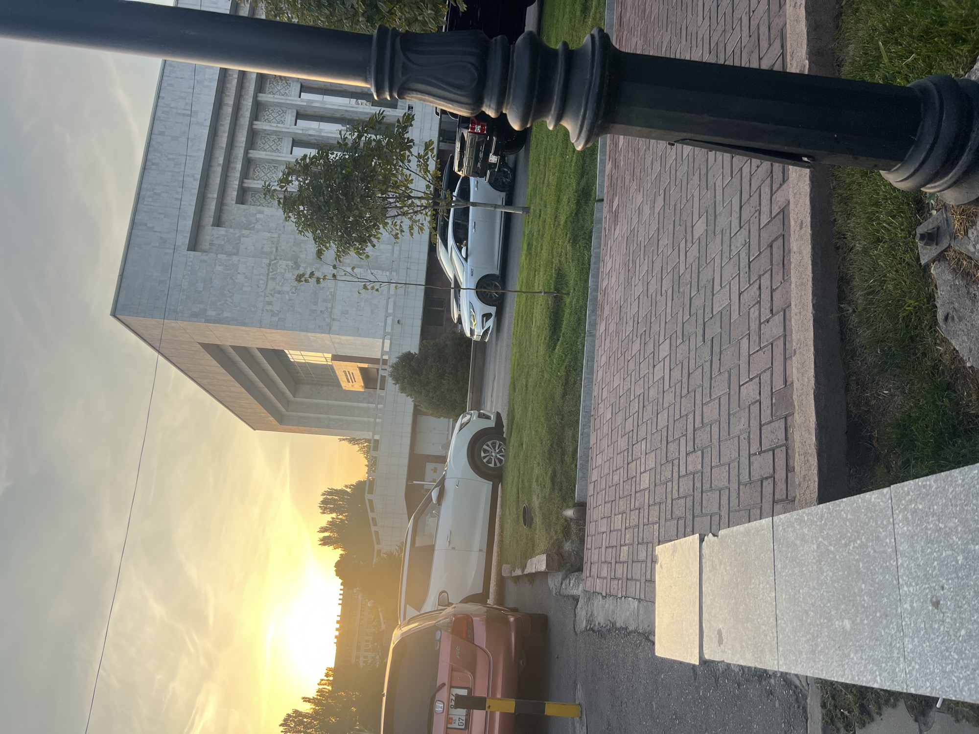 The image captures a street scene in Bishkek, Kyrgyzstan, during what appears to be sunset. In the background, a large, modern building with a light-colored, textured facade dominates the skyline. The building features a grand entrance with tall, narrow windows and decorative elements. To the left of the building, a row of trees is silhouetted against the bright orange and yellow hues of the setting sun. The sky displays a gradient of colors, from pale yellow near the horizon to a lighter gray higher up, with some wispy clouds visible.
In the foreground, several vehicles are parked on the side of the road and on a grassy area. A red hatchback is parked on the left, its rear end visible. Next to it, a white SUV is parked on the grass. Further down the street, a white sedan is parked on the asphalt, and behind it, a dark-colored pickup truck. The road is paved with asphalt, and a pedestrian walkway made of red brick pavers is situated in the lower right portion of the image. A black lamppost with ornate detailing stands prominently in the right foreground, partially obscuring the view of the street and building. The ground is a mix of grass, pavement, and some concrete borders. The overall impression is a quiet urban setting bathed in the warm light of sunset.