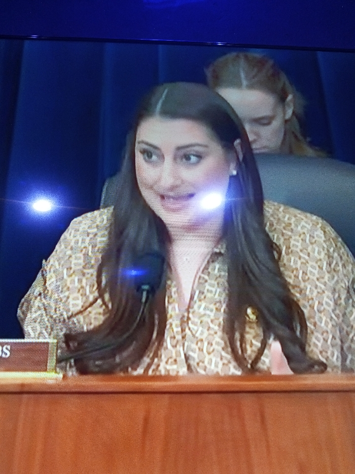 A brunette woman with dark hair parted in the middle is speaking into a microphone at a desk. She wears a patterned tan and white blouse. Behind her is another woman with light brown hair and a dark gray chair. The backdrop is a dark blue color. A light flare shines near the woman's right eye.