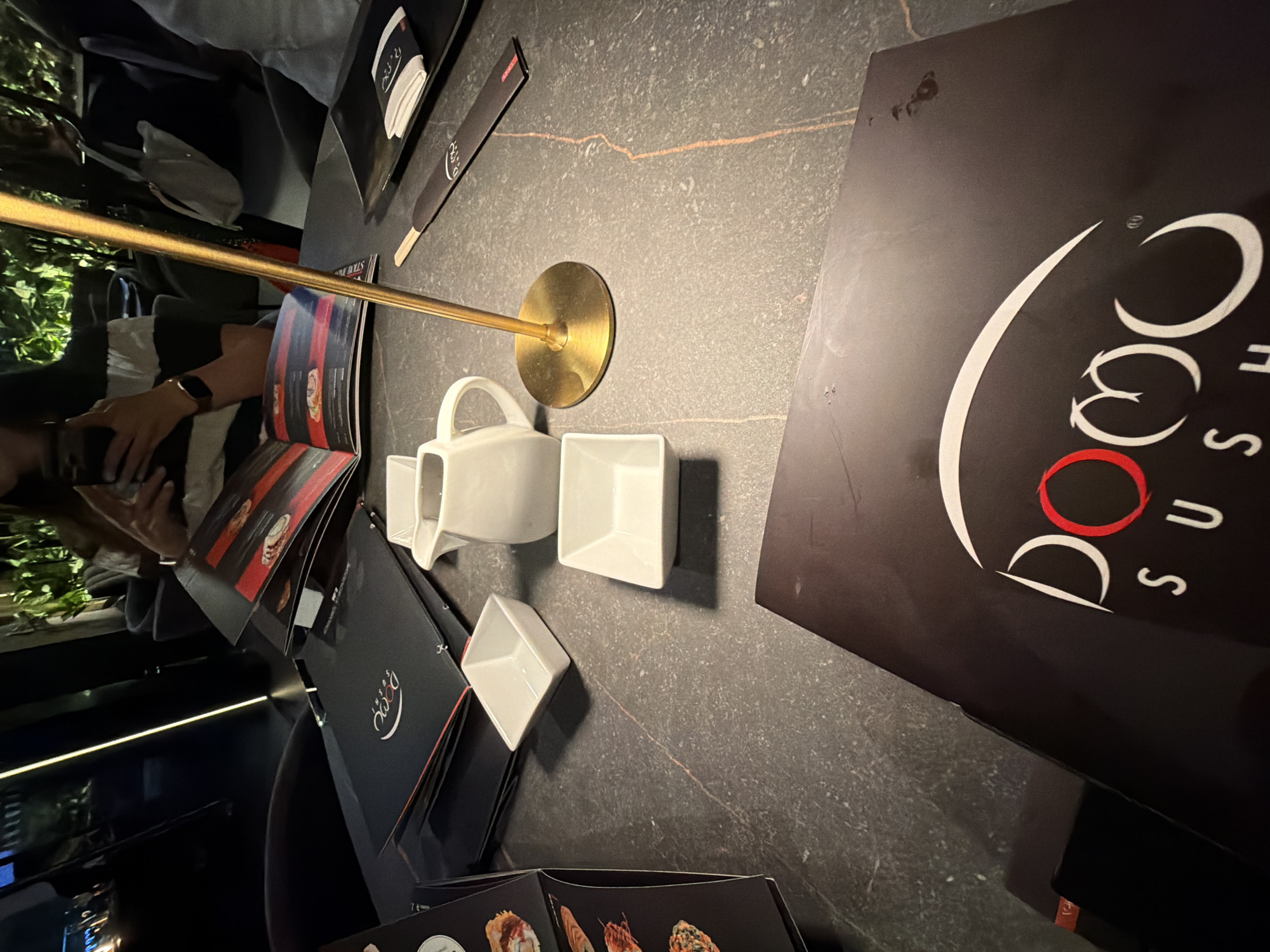 The image depicts a table at what appears to be a restaurant called "Domo Sushi." The table surface is dark and textured. A golden-colored adjustable lamp stands on the table. There is a white teapot and two small square white dishes. Menus are present, along with chopsticks in a paper wrapper. A person is partially visible in the background. The location is likely San Bernardino Tlaxcalancingo, Mexico, based on the reference to Domo Sushi.