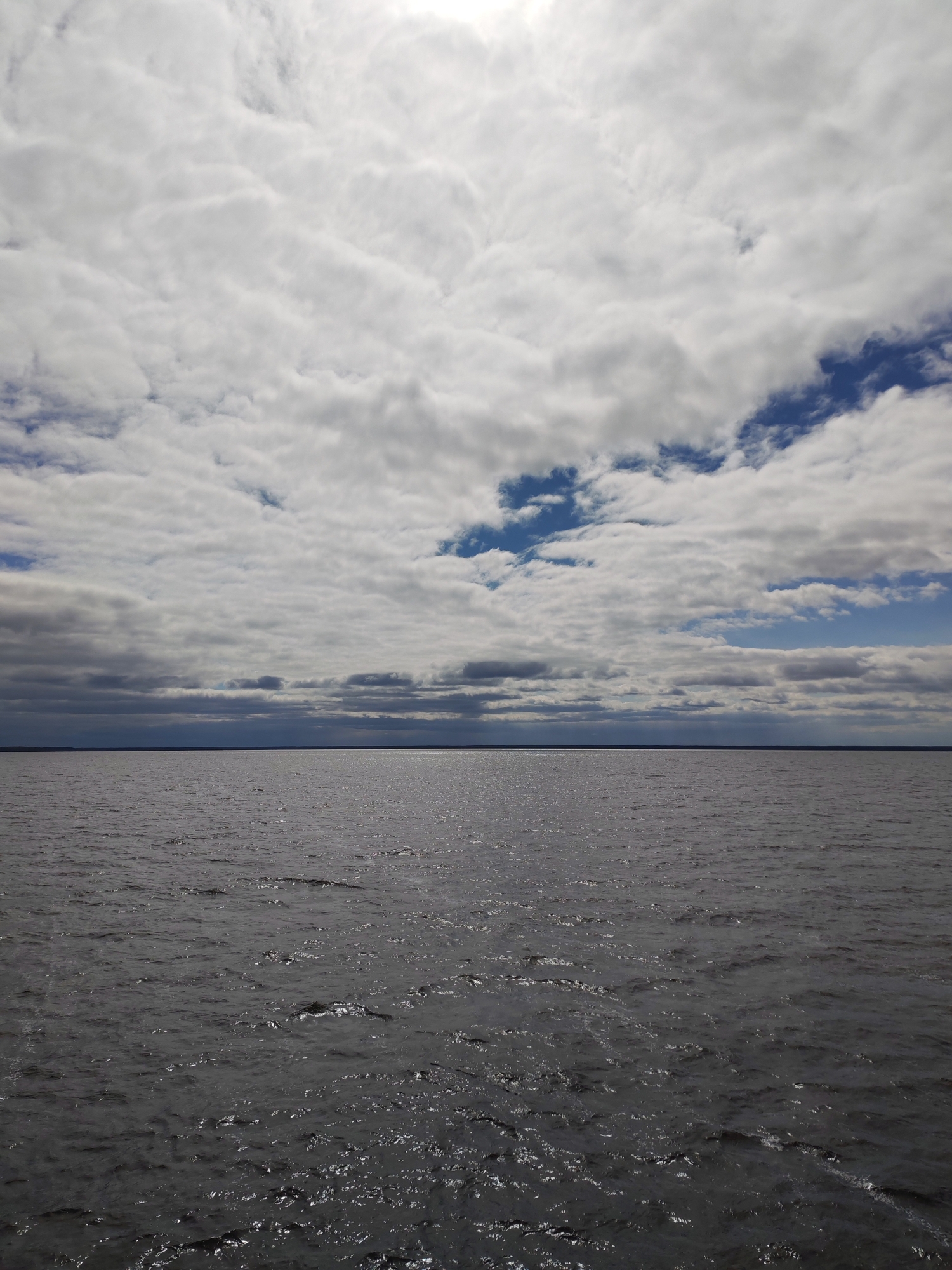 The image presents a wide, expansive view of a large body of water under a dynamic sky, located in Pochinok, Russia. No people or specific objects are visible, emphasizing the natural landscape.
The upper half of the frame is dominated by a sky filled with layered, mostly white and light grey clouds. These clouds appear to be stratocumulus or altocumulus formations, with some brighter areas suggesting the presence of sunlight behind or above them. Through breaks in the cloud cover, particularly towards the right and center-left, patches of clear blue sky are visible.
A distinct, flat horizon line cleanly separates the cloudy sky from the water below. There's a very faint, dark line just above the horizon on the water side, which could indicate a distant landmass or shoreline.
The lower half of the image features a vast expanse of dark grey water, likely a lake or a wide river. The water's surface is not entirely smooth, displaying numerous small ripples and gentle waves. These ripples catch and reflect the ambient light, creating a shimmering, textured effect across the otherwise dark water. The scene conveys a sense of serene vastness and natural grandeur, captured during the daytime under variable cloud conditions. There is no visible text in the image.