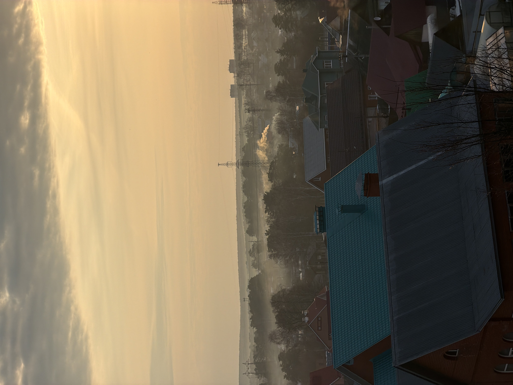 The image shows a hazy, early morning scene in Chaikovsky, Russia. In the foreground are residential buildings with distinct roof colors: blue-green and dark grey. Beyond the houses, the view opens to a flat landscape shrouded in a light mist. Transmission towers are visible in the distance, suggesting power lines running through the area. There is a hint of smoke or steam rising in the distance. The sky is a hazy yellow-gold color.
