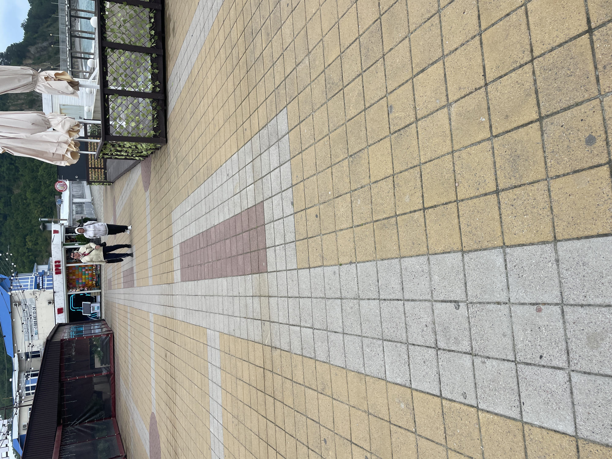 The image shows a paved walkway in Olginka, Russia. The walkway is made of square tiles in alternating colors of yellow, grey, and reddish-brown. Two people stand on the walkway. One is wearing a white jacket and the other a beige jacket. The walkway is bordered by buildings and vegetation on the left side. On the right side is a view of a beach with a metal railing. The sky is overcast.