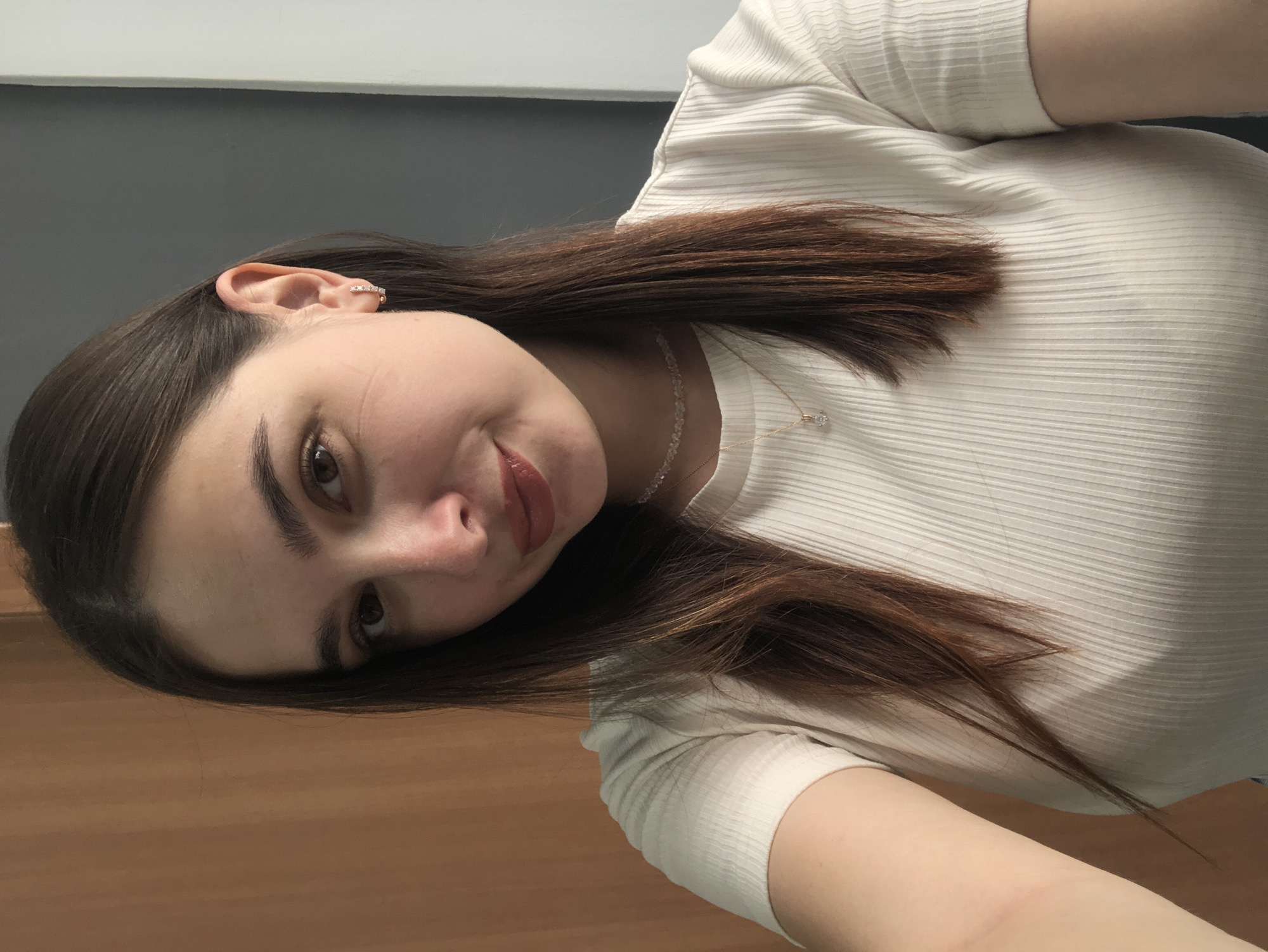 This is a close-up, head-and-shoulders portrait of a young woman with long, dark brown hair. She is wearing a white, ribbed shirt and a delicate necklace with a small pendant. Her head is tilted, and she is looking towards the camera with a slight smile. She has dark eyebrows, brown eyes, and is wearing lipstick. A diamond stud earring is visible in her left ear, and a decorative ear cuff with small stones is also present on the same ear. The background is blurred, showing a grey wall and a wooden surface in the foreground, suggesting an indoor setting, possibly a desk or table. The lighting appears to be natural or soft indoor lighting. No text is visible in the image. The overall mood is relaxed and casual. The location context is Chaikovsky, Russia, but there are no specific visual cues to confirm this.