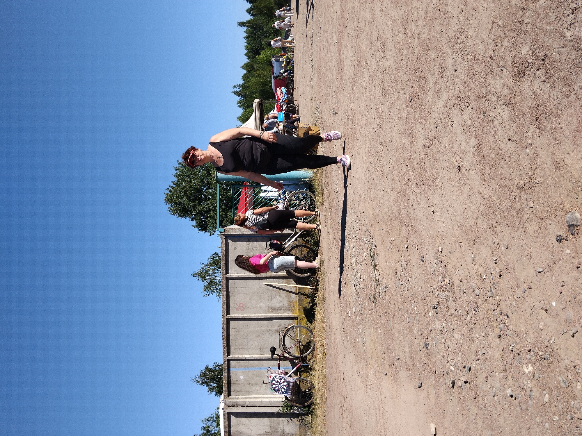 A medium shot captures an outdoor scene with a clear blue sky and a dusty, gravel-strewn ground. In the foreground, a woman wearing a black tank top, black leggings, and pink and white sneakers walks towards the camera. She has short brown hair and is wearing a red visor. A black fanny pack is around her waist. Behind her, to the left, is a concrete wall. Two bicycles are leaning against the wall. One bicycle on the far left has a dartboard attached to its frame. Two women are standing near the bicycles, one wearing a pink t-shirt and denim shorts, the other wearing a black and white striped shirt and black shorts. Further in the background, there are several market stalls with tents and vehicles, and a few more people are visible. The scene appears to be a market or an outdoor event.