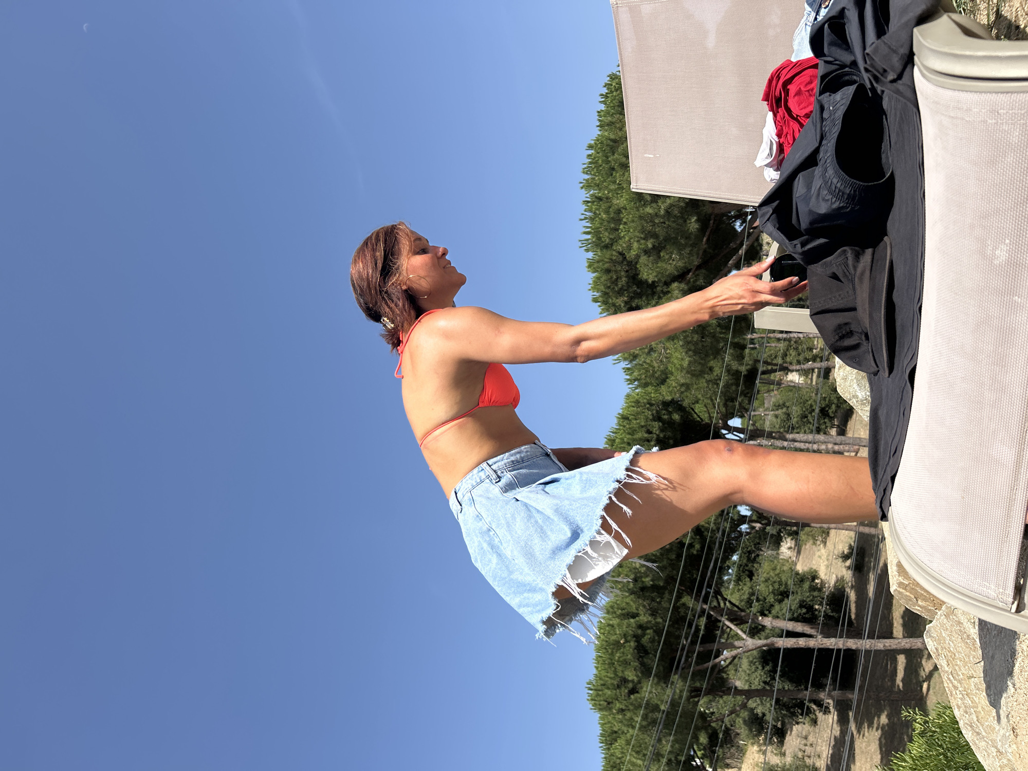 A woman in an orange bikini top and a denim skirt with frayed edges bends over a lounge chair outdoors. She is reaching for a pile of dark clothing on the chair. The background shows a clear blue sky, green trees, and electrical wires. The scene appears to be in Sant Vicenç De Montalt, Spain, near a residential area, indicated by the landscape and trees.