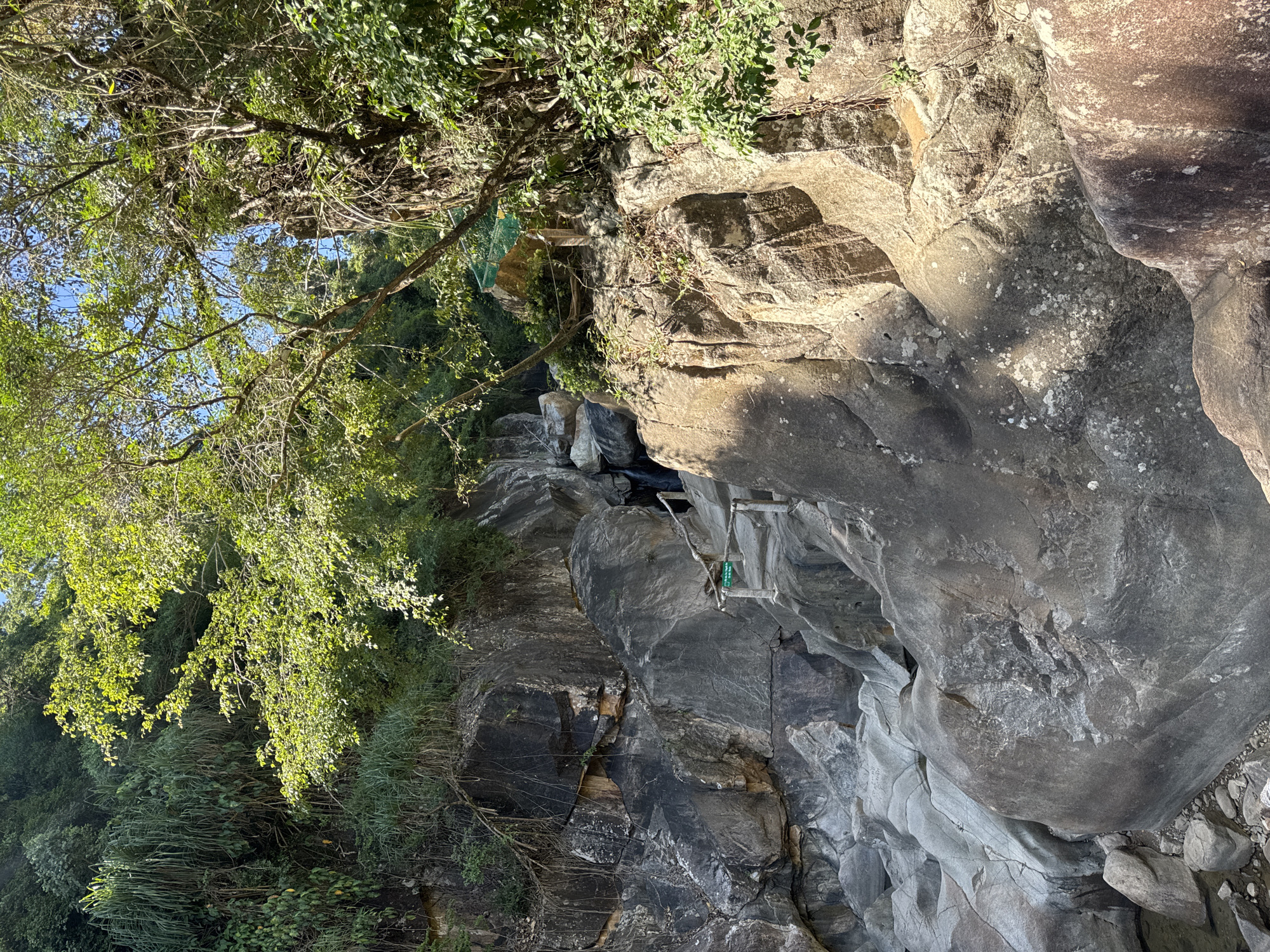 The image depicts a rocky incline with vegetation and a small waterfall in the distance, located near the city of Ninh Hoa, Vietnam. The scene is dominated by large, weathered rocks of varying shades of gray and brown, interspersed with lush green foliage. Sunlight filters through the trees, casting dappled shadows on the rocks. A wooden railing with a green sign is visible on the left side of the frame, suggesting a pathway or viewing area. The waterfall, partially obscured by rocks and trees, appears to be a small cascade. The overall impression is a natural, somewhat rugged environment, possibly a tourist destination or a natural landmark. There are no people visible in the image.