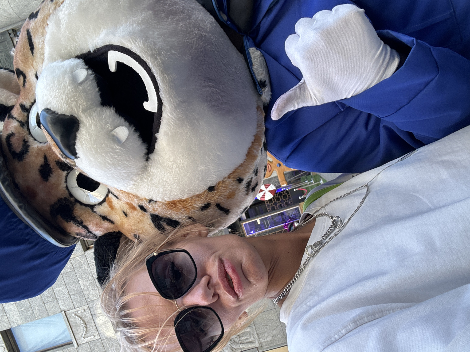 A woman with blonde hair and sunglasses is taking a selfie with a mascot of a cheetah wearing a blue hat and jacket. The woman is wearing a white t-shirt and a silver necklace. The mascot is wearing white gloves and giving a thumbs-up. The background shows a building with gray stone walls and a blue window. The scene appears to be outdoors in a city environment. There is no specific information indicating the location is in Saint Petersburg, Russia.