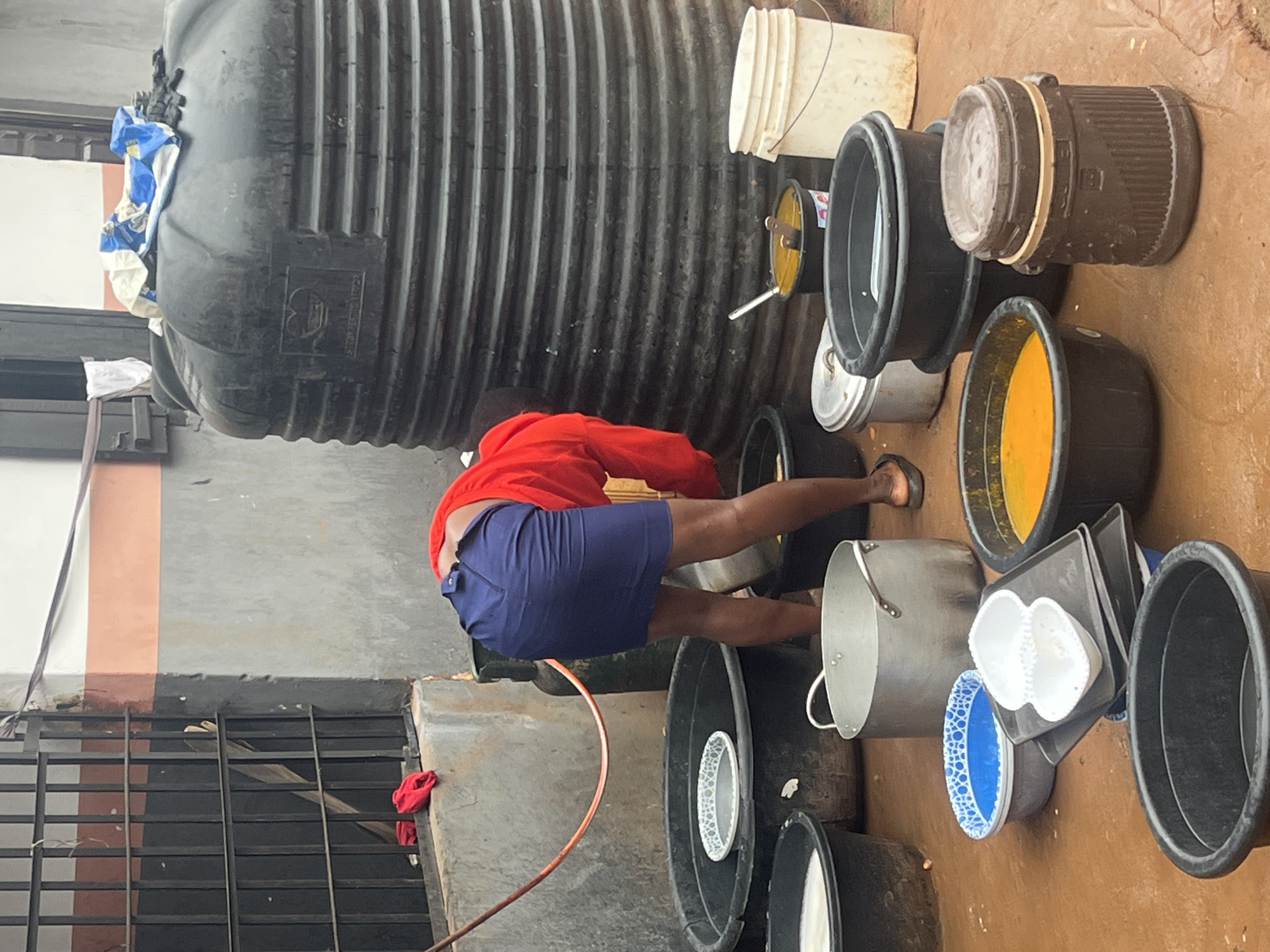 In Nkpor, Nigeria, a person wearing a red shirt and a blue skirt is bending over, appearing to be engaged in some task outdoors. The scene is characterized by a collection of large black plastic tubs, a sizable black water tank with ribbed sides, and a large aluminum pot with handles. Several smaller containers, including a blue and white patterned basin, white plastic containers, and a dark brown cylindrical container with a lid, are also present. A dark metal grate is visible in the background, along with a concrete wall painted with horizontal stripes of peach and gray. The ground is a reddish-brown, muddy surface. A red hose is draped over the grate and extends towards the tubs.