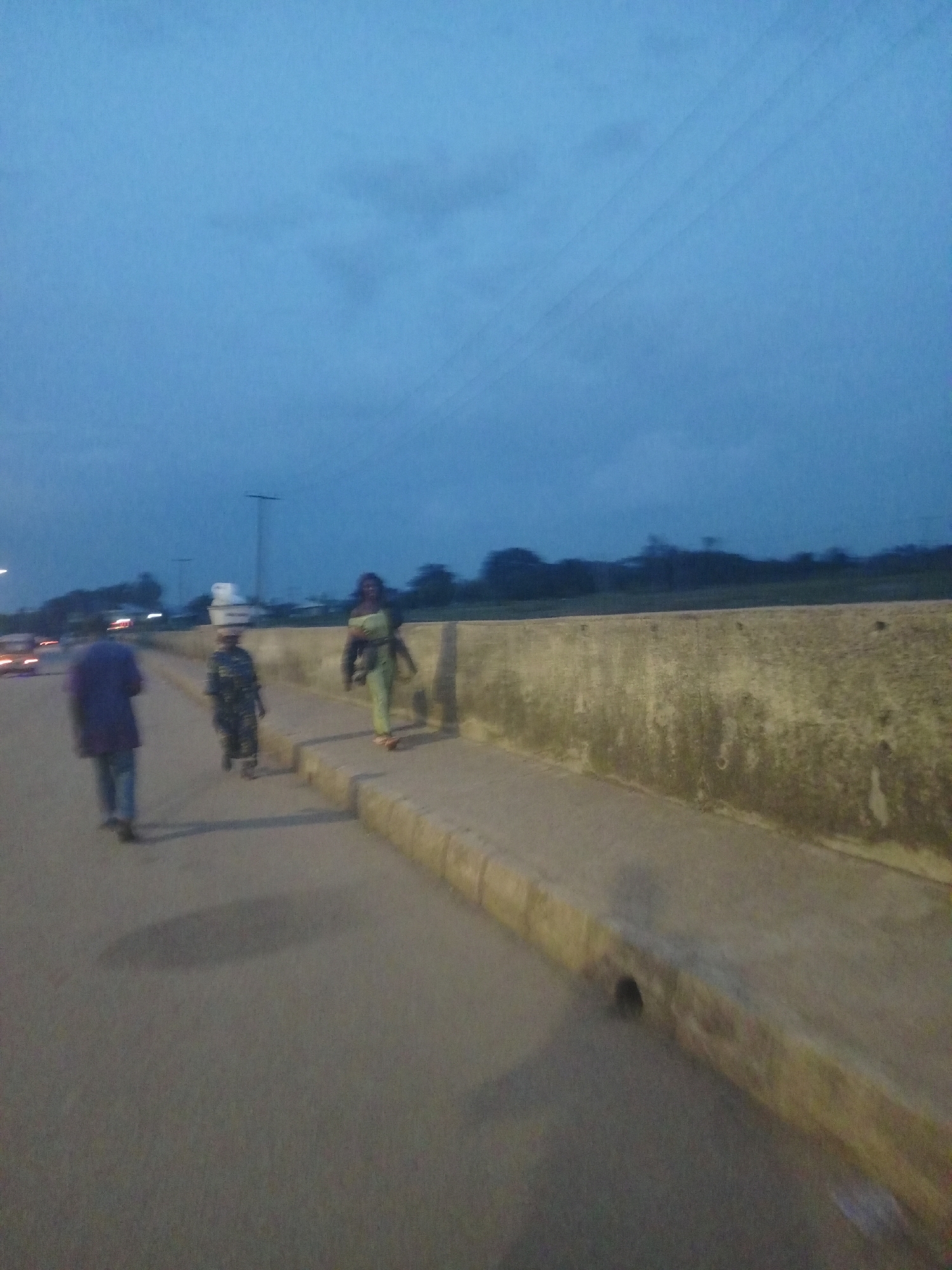 The image depicts a twilight scene on a paved road, likely in Bukuru, Nigeria, with three individuals walking. The sky is a deep blue with scattered clouds. On the left side of the road, a person wearing a dark blue top and blue trousers walks away from the camera. Further down the road, a person in a camouflage-patterned outfit carries a white object on their head. To the right of this individual, another person wearing a light green garment walks towards the camera. The road is bordered by a concrete barrier, and a storm drain is visible at the edge of the pavement. In the background, trees and vegetation are silhouetted against the sky, and distant lights suggest an urban environment. The overall atmosphere is subdued, consistent with dusk.