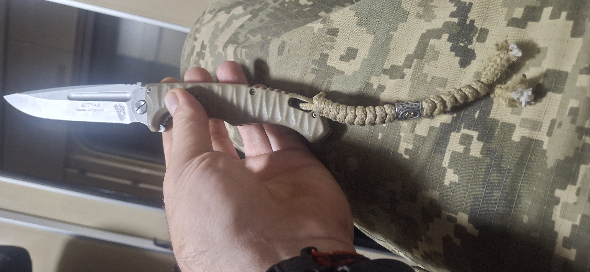 A person's hand holds a tan-handled folding knife. The blade is partially open and made of polished metal. Engraved text on the blade appears to read "KTTAP" and "HOK". A braided tan cord is attached to the knife's pommel and hangs down, featuring a decorative metal bead with a silver Celtic-like design and a golden element. The person is wearing camouflage pants. The background is out of focus but suggests an indoor setting with vertical elements. The location is identified as Orlivschyna, Ukraine.