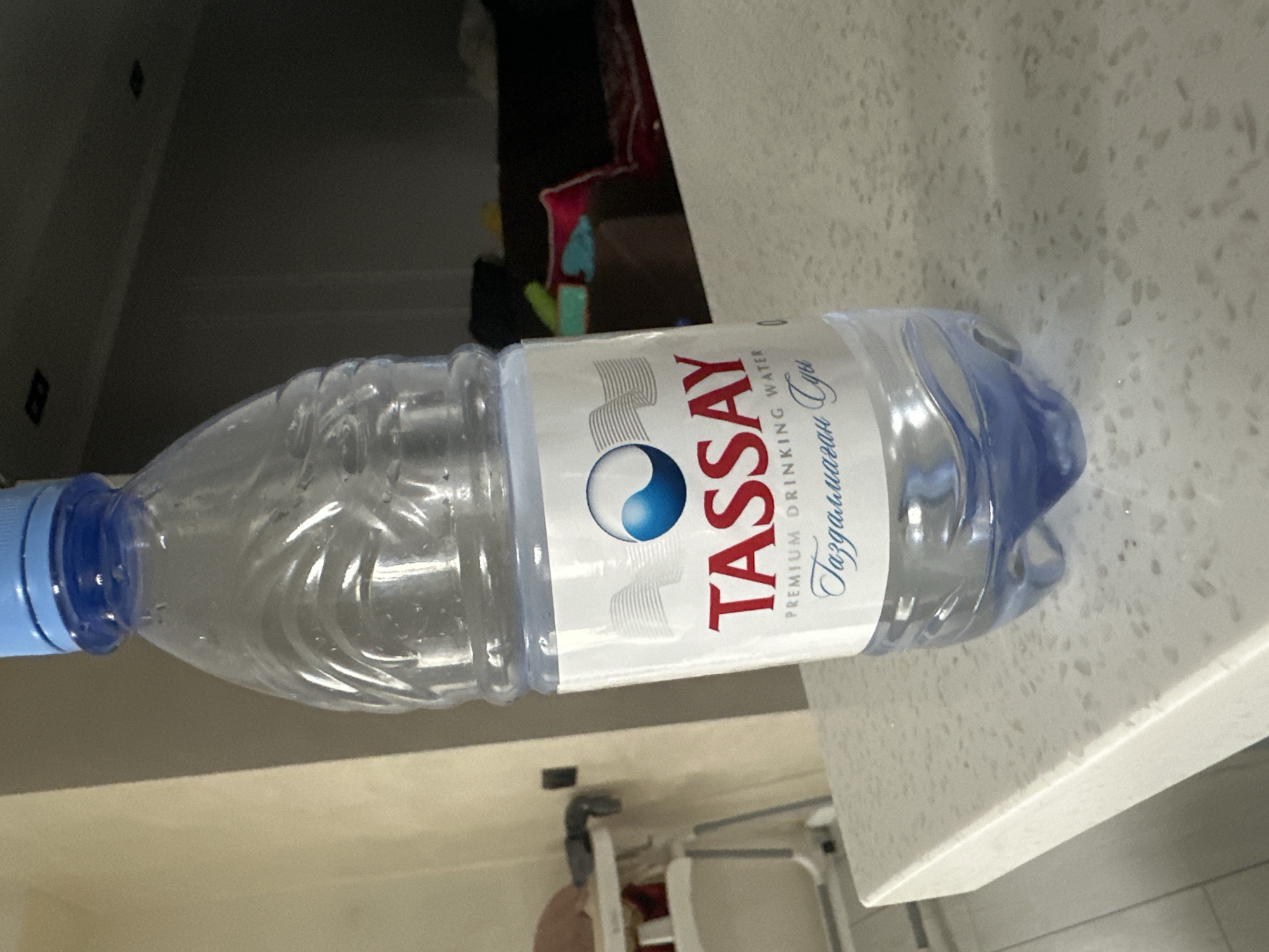 A clear plastic bottle of Tassay Premium Drinking Water, with a blue cap, is centered in the foreground, standing on a speckled white countertop. The bottle's label is white with red text that reads "TASSAY" and smaller text below that says "PREMIUM DRINKING WATER" and then text in a script font in what appears to be Kazakh. A blue and white circular logo is on the label. The background is out of focus and shows a blurred interior environment, possibly a home, with furniture and a wall. There is no discernible location in the city of Almaty, Kazakhstan.