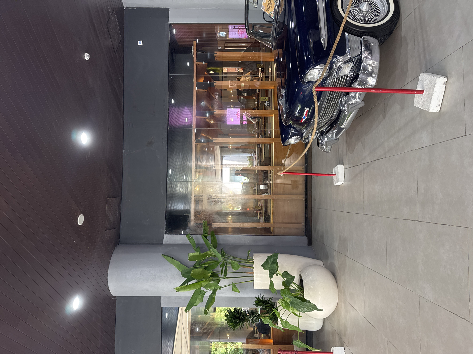 The photograph depicts the interior of a commercial space, potentially a restaurant or bar, located in an unidentified area of Unknown, Unknown. A classic blue car, possibly a vintage Chevrolet, is displayed as a decorative element near the entrance or a prominent area. The car is roped off by red stanchions with white bases, suggesting it is not to be touched or entered.
To the left of the car, a large glass wall reveals an interior space with wooden shelving and decor, with a glimpse of patrons inside. A dark grey column supports the structure, adjacent to which are planters with leafy green plants, adding a touch of nature to the indoor environment. The flooring is a light grey tile, and the ceiling is a dark brown wood paneling with recessed lighting. The lighting appears to be a mix of natural light from the outside and artificial light from within the establishment. The overall atmosphere is modern with classic elements. No people are clearly visible in the immediate foreground, but figures can be discerned in the interior space behind the glass wall.