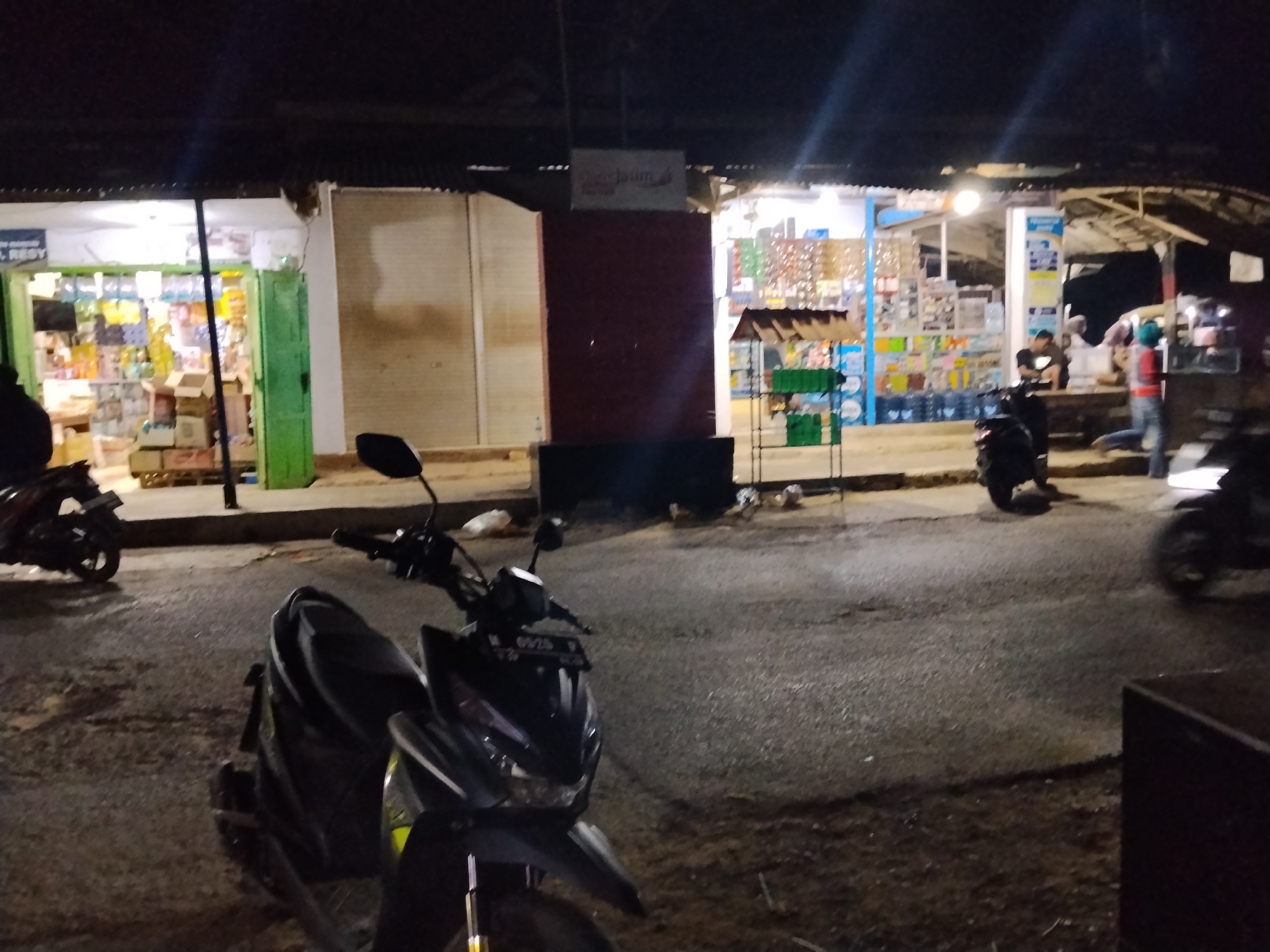 In Arjasa, Indonesia, at night, a street scene unfolds in front of a row of small shops. A dark-colored scooter is parked in the foreground, angled towards the viewer, its front section visible. Further down the street, another dark-colored scooter is parked on the left side. In the middle ground, a person wearing a red shirt and blue jeans stands near a shop. Another individual, partially obscured, appears to be seated or leaning against a structure behind the parked scooters. The shops themselves have open fronts displaying various goods, including bottled drinks and packaged items, illuminated by overhead lighting. A large, maroon-colored rectangular structure stands between two of the shops. The street surface is asphalt and appears wet or reflective, suggesting recent rain or the presence of water. The overall lighting is dim, characteristic of nighttime.