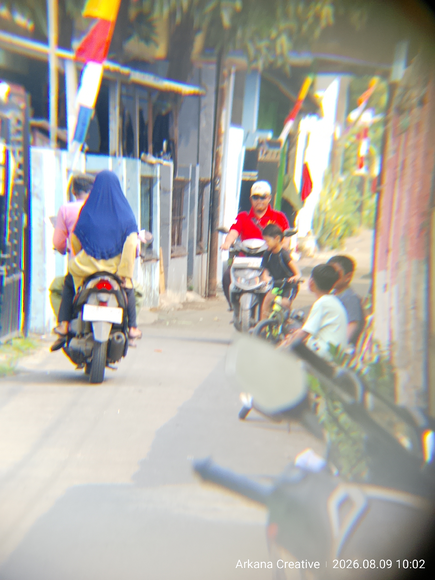 In Bekasi, Indonesia, on August 9, 2026, at 10:02 AM, a street scene unfolds with multiple individuals and vehicles. A person in a red shirt and a white cap is riding a motorcycle, accompanied by a child in a black shirt behind them. To the left, another motorcycle is being ridden by a person wearing a blue headscarf and a yellow outfit, with another person visible behind them wearing a pink shirt. Two young boys on a bicycle are also present in the scene, with one wearing a white shirt and the other a grey shirt. The backdrop consists of buildings with flags and foliage, suggesting an urban environment. The image is characterized by a noticeable chromatic aberration and a lack of sharp focus, giving it a somewhat abstract appearance.