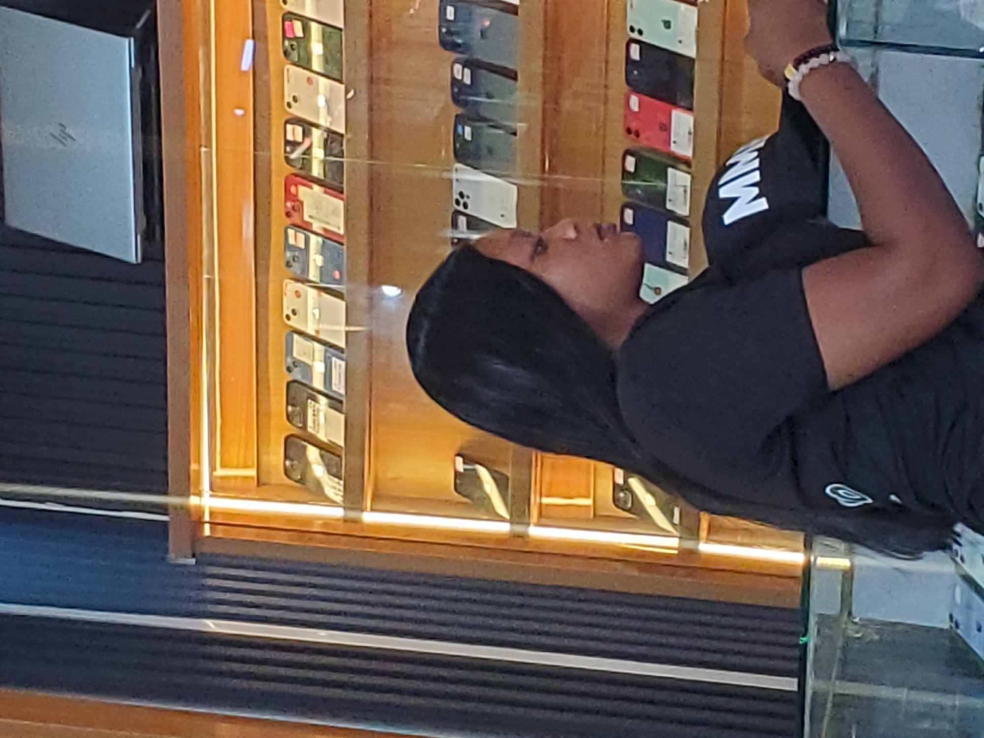 The image shows a close-up of a person, likely a woman, in a retail setting. She has long black hair and is wearing a black t-shirt with white lettering on the sleeve, which appears to say "MW". She is also wearing a beaded bracelet on her wrist. The background suggests a display case filled with mobile phones. The phones are arranged in rows, and many are visible in their packaging, showcasing different colors like white, red, green, and blue. Some phone cases are also visible. To the left of the phone display is a light-colored laptop or tablet device. The lighting is bright and seems to come from within the display case, creating reflections on the glass. The overall scene appears to be an indoor electronics store. The context of Jos, Nigeria, suggests this is likely a phone shop within the city. The image's orientation is rotated, with the person appearing to be looking to the right.
