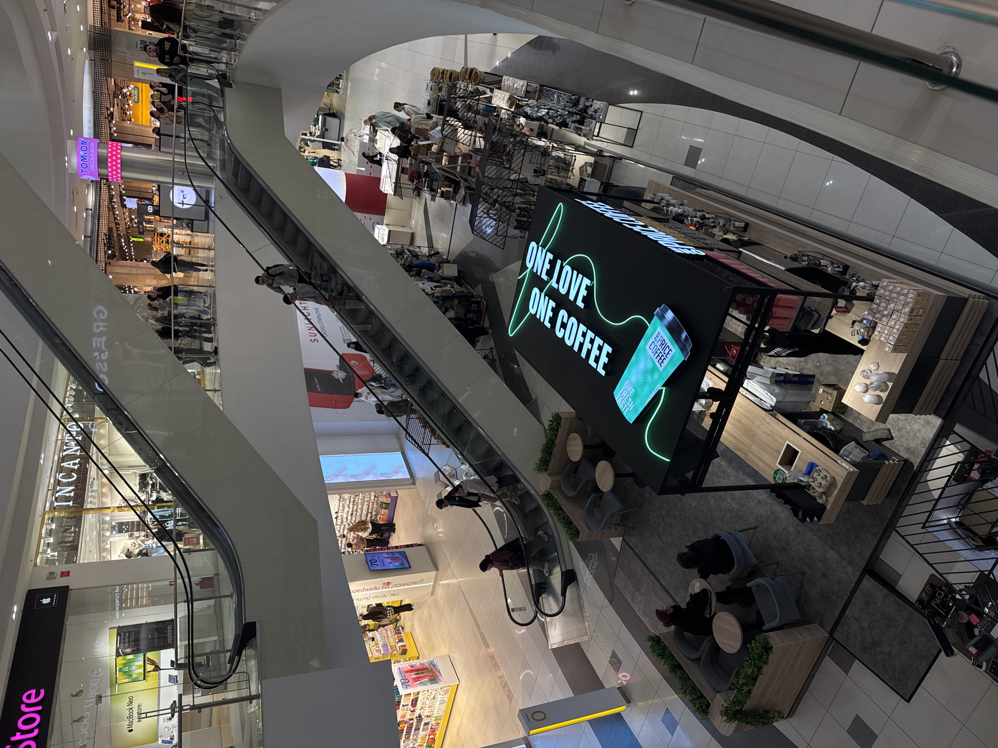 An elevated, diagonal shot captures the vibrant interior of a multi-level shopping mall. Several escalators diagonally traverse the frame, connecting various floors, with numerous people visible on them and strolling along the different levels. The mall appears modern, with bright lighting and polished surfaces.
In the center-right, a prominent coffee shop is visible, featuring a large, illuminated sign that reads "ONE LOVE ONE COFFEE." A graphic of a coffee cup on the sign also includes text like "PRICE COFFEE TASTE FRESH." Around the coffee shop, some tables and chairs are arranged, with a few people seated, enjoying beverages. The counter of the coffee shop displays various items.
Storefronts line the upper and lower levels. Notable brands and text include "INCANTO" on an upper-level store, and "SUNLIGHT" appearing multiple times on signs, suggesting a jewelry or accessory store. Another sign reads "TOMTOM BUBBLE CAFE." On a lower level, there's an "iStore" with text like "MacBook Neo" and "restore." Other visible Russian text includes "так легко быть улыбнись ты прекрасн" (it's so easy to be, smile, you are beautiful) and "СКОРО ОТКРЫТИЕ" (Coming Soon), along with "БАДЫ" (Supplements). Discount signs for "50%" and "70%" are also visible.
The scene depicts typical mall activity, with shoppers moving between levels, browsing stores, and patronizing the coffee shop. The architecture suggests a spacious and contemporary commercial environment.