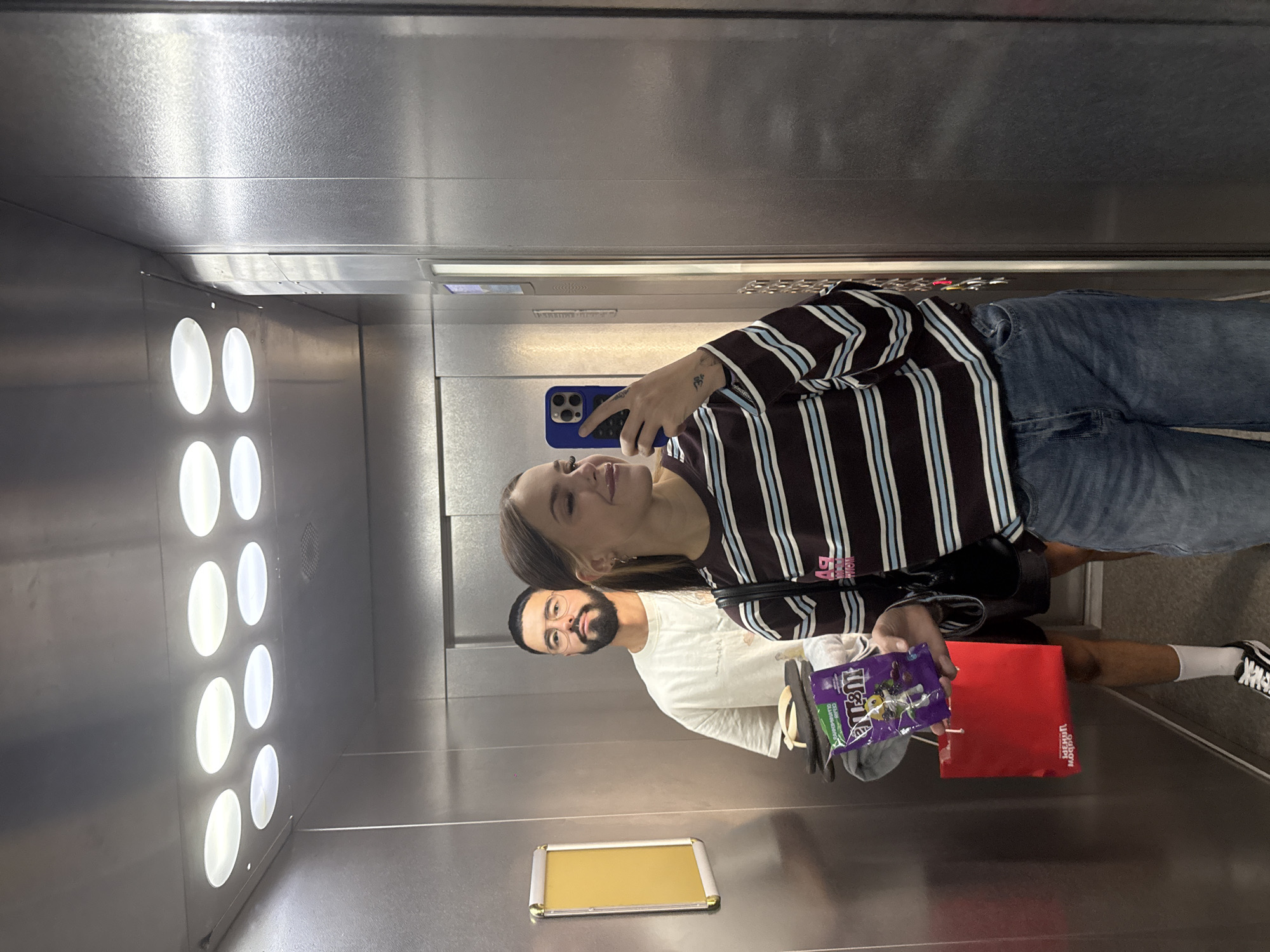 Two individuals are taking a selfie inside a stainless steel elevator. The woman in the foreground, holding a smartphone with a blue case, is wearing a striped long-sleeved shirt and denim jeans. She has blonde hair and is looking towards the phone. A man is partially visible behind her, wearing a white t-shirt and glasses, with a beard. He is holding a red bag and a package of M&M's. The elevator has a mirrored ceiling with circular lights and stainless steel walls. The image appears to have been taken in Chelyabinsk, Russia.