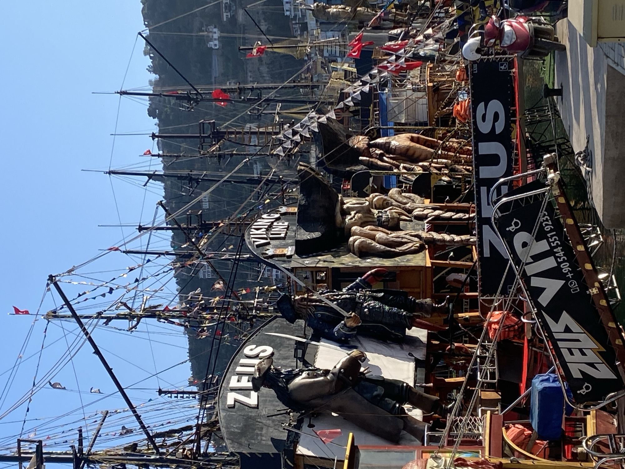 This image captures a vibrant, elaborately decorated pirate-themed tour boat, likely docked in a harbor under a clear blue sky, in Alanya, Turkey. The multi-masted vessel is adorned with numerous statues and figures, including several pirates wielding swords, a large eagle or bird sculpture on the bow, and other monstrous or mythological figures resembling a kraken or sea creature.
The boat's rigging is complex and festooned with strings of triangular bunting and various flags, including the red Turkish flag with its white crescent and star, and several black pirate flags. Prominently displayed on the side of the boat and on large banners are the words "ZEUS" and "VIP," indicating its name and a potential service tier. One banner also shows "VIKING LINE" and a Turkish phone number: "+90 544 551 9239," alongside accessibility icons. Orange life rafts or buoys are visible on deck, indicating safety provisions.
The setting suggests a bright, sunny day, with rugged, tree-covered hills or mountains forming the backdrop. The overall scene conveys a lively, tourist-focused atmosphere, common in popular coastal destinations. The boat itself appears to be a major attraction, designed to offer a themed experience to its passengers.