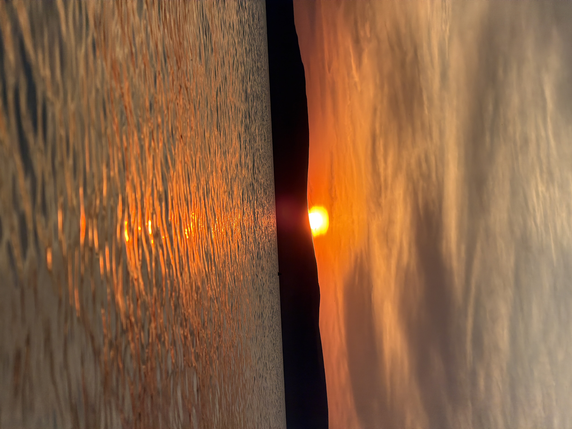 A vertical, full-frame shot captures a sunset over a body of water, with a dark, silhouetted hill in the background. The sun, a bright orange orb, is partially obscured by the hill, casting a warm glow across the sky and reflecting onto the rippling water. The sky is filled with wispy clouds in shades of orange, yellow, and grey, indicating the transition from day to night. The water is calm, with gentle waves reflecting the sunset's colors. The scene appears serene and tranquil.