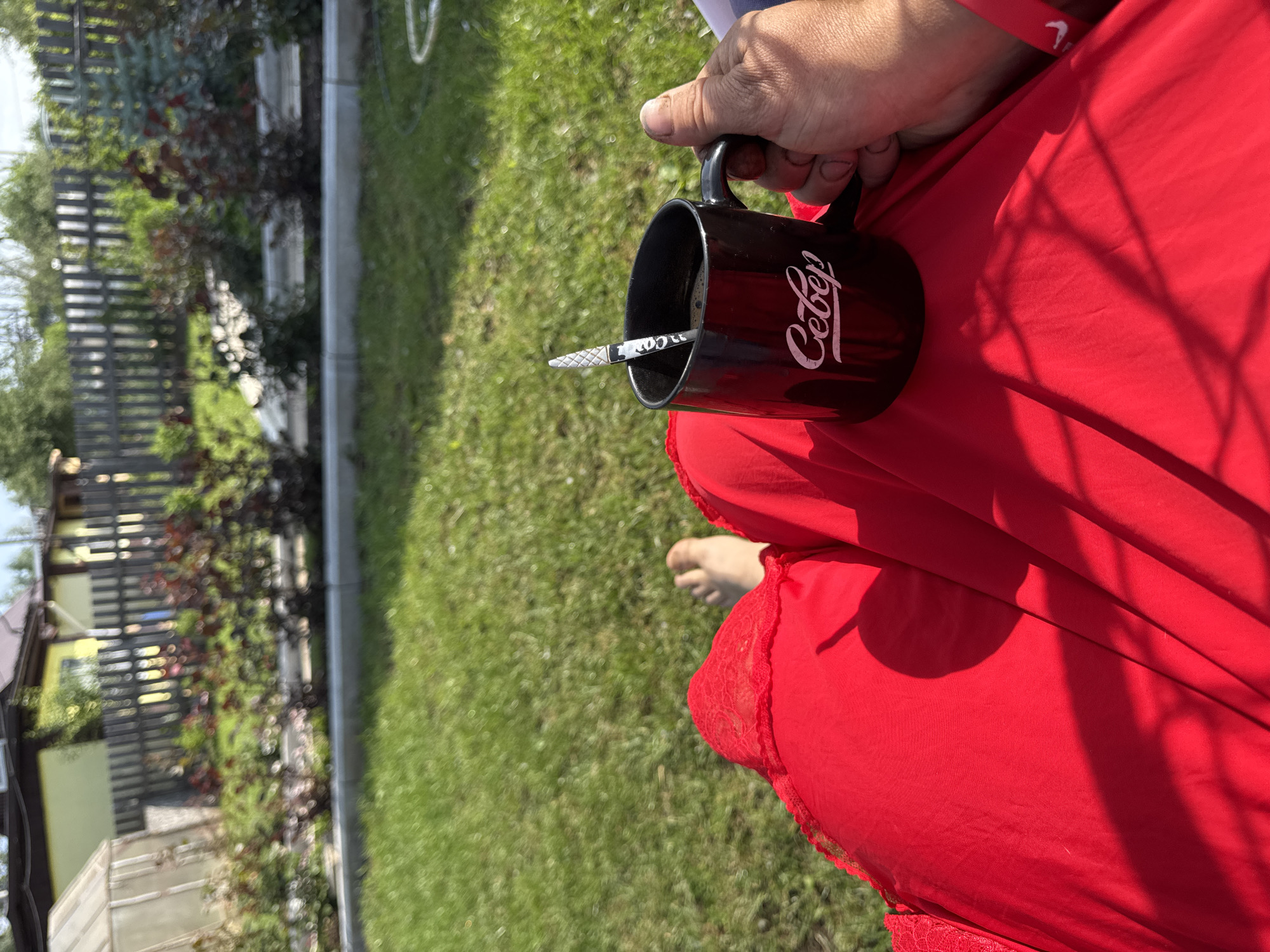 The image shows a person relaxing in a grassy backyard, holding a black mug with the word "Север" (Sever) written on it in white script. The person is wearing a bright red garment, possibly a dress or skirt, with lace trim visible at the top. Bare feet are visible on the grass. In the background, a wooden fence separates the yard from a garden with red-leafed bushes and a greenhouse. Beyond the fence, a yellow house with a brown roof is partially visible. The scene appears to be set in a residential area on a sunny day.