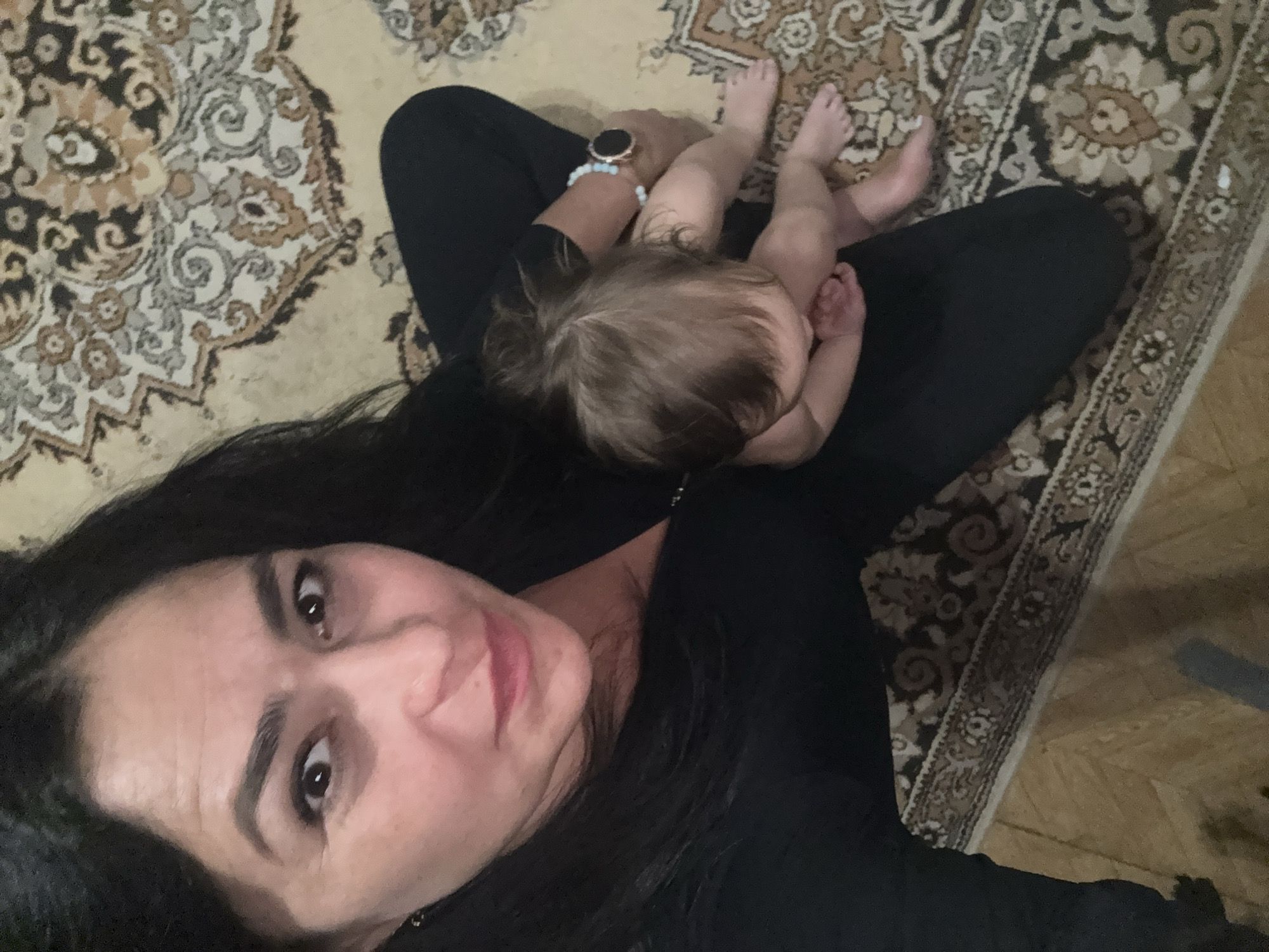 An indoor, overhead selfie captures a woman and a baby lying on a patterned carpet. The woman, with dark hair and a neutral to slightly smiling expression, looks directly at the camera. She wears a black top and a smartwatch with a light blue beaded bracelet on her right wrist. An infant with light brown hair is nestled on her lap or chest, visible from the back and side, showing small legs and feet.
The subjects are positioned on a traditional-style carpet with intricate floral and geometric patterns in shades of beige, brown, grey, and black. A small portion of a light-colored wooden floor is visible at the bottom right of the frame, suggesting the carpet covers only part of the room. The lighting appears soft and internal. The activity depicts a tender, intimate moment between what appears to be a mother and child, possibly resting or cuddling. No text is visible in the image. The scene is set in Chaikovsky, Russia.