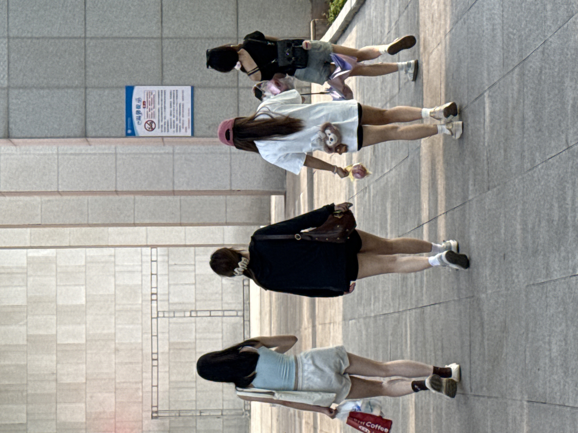 A medium shot captures four young women walking along a sidewalk on a bright, sunny day. They appear to be in Jiangjin, China, based on signage in the background. The women are all dressed casually in summer attire and are walking in the same direction. The first girl wears a light blue shirt with short sleeves, a pair of white short, white socks, and black shoes. She also carries a paper bag and a white cloth. The second girl wears a dark jacket with short sleeves, a pair of short, white socks, and white shoes. She also carries a brown bag. The third girl wears a white shirt with a toy bear and a pink hat with short sleeves, a pair of short, white socks, and white shoes. She also carries a toy. The fourth girl wears a black shirt with short sleeves, a pair of short, white socks, and brown shoes. She also carries a black bag. The sidewalk is made of gray stone and there is a building to the left. The building is made of gray stone and has a blue and white sign on the wall. The background is out of focus, but it appears to be a busy street with people and cars.