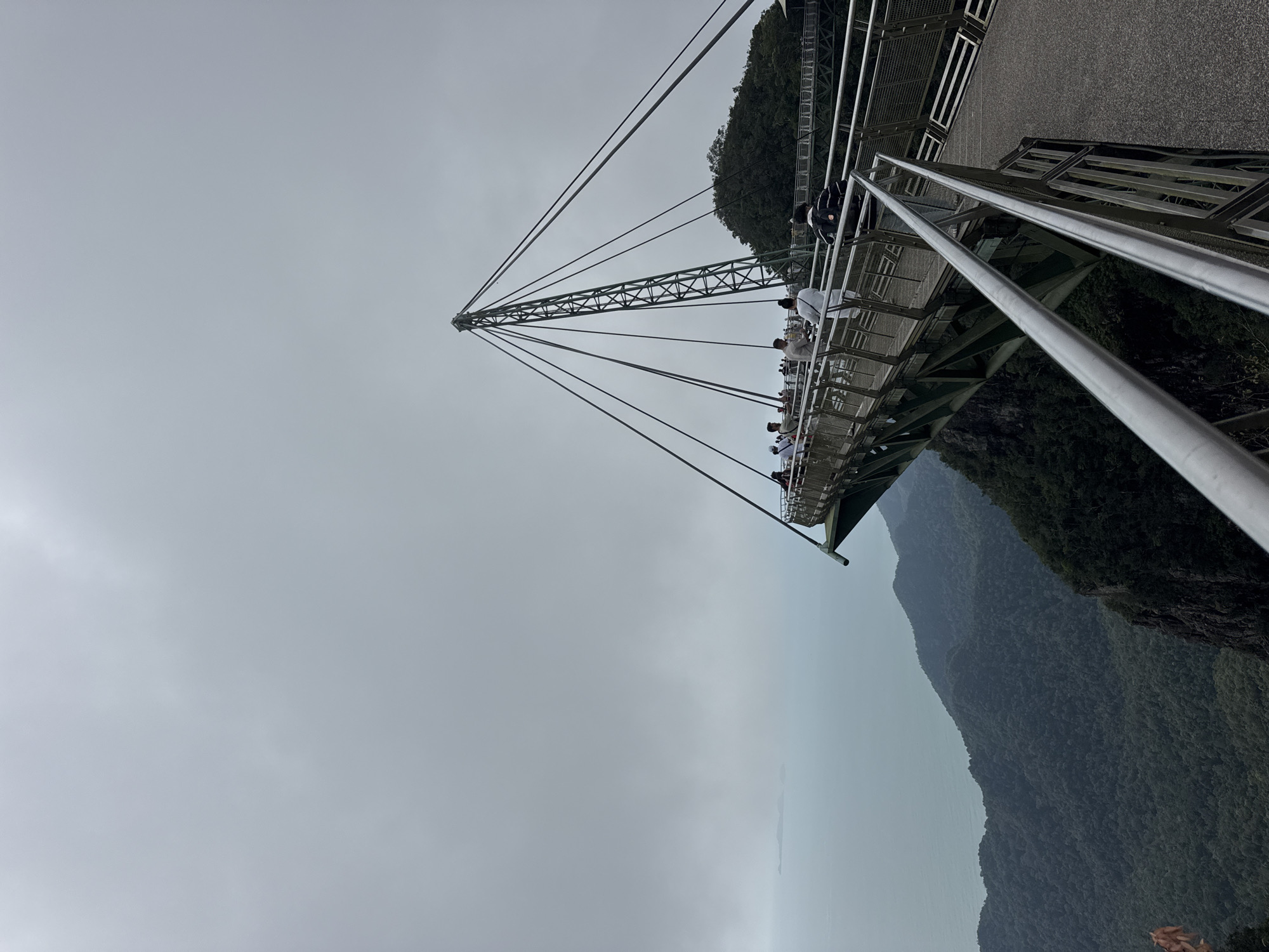 The image depicts the Langkawi Sky Bridge in Malaysia, a pedestrian walkway situated high above the rainforest. The bridge is characterized by its curved design and the prominent suspension tower with numerous cables. A cloudy, overcast sky dominates the upper portion of the frame. Below the bridge, dense, green, mountainous terrain stretches towards the ocean, which is visible in the distance with a faint outline of islands. Several people are present on the bridge, some walking and others observing the view. The bridge itself is constructed with metal railings and a paved walkway. The viewpoint is from the bridge, looking outwards and downwards.