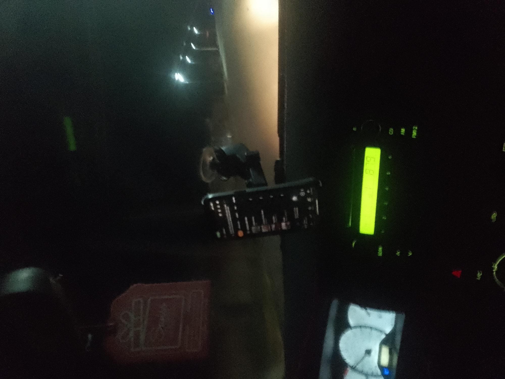The image depicts the interior of a vehicle at night. A smartphone is mounted on a car dashboard using a black suction cup holder. The phone's screen displays an application with vertical bars, suggesting music playback or a similar visualizer. To the right of the phone, a car radio display is visible, showing the number "879" in green digital font. The radio also has several buttons and indicators, including "CD", "AM", and "FM1/2". Below the radio, a portion of a car's instrument cluster is visible, featuring gauges with needles. In the foreground, to the left of the phone, a red keychain with a white emblem is hanging. The overall lighting is dim, consistent with a nighttime interior shot inside a car. The background is dark and out of focus, with some indistinct lights and shapes that are not clearly identifiable.