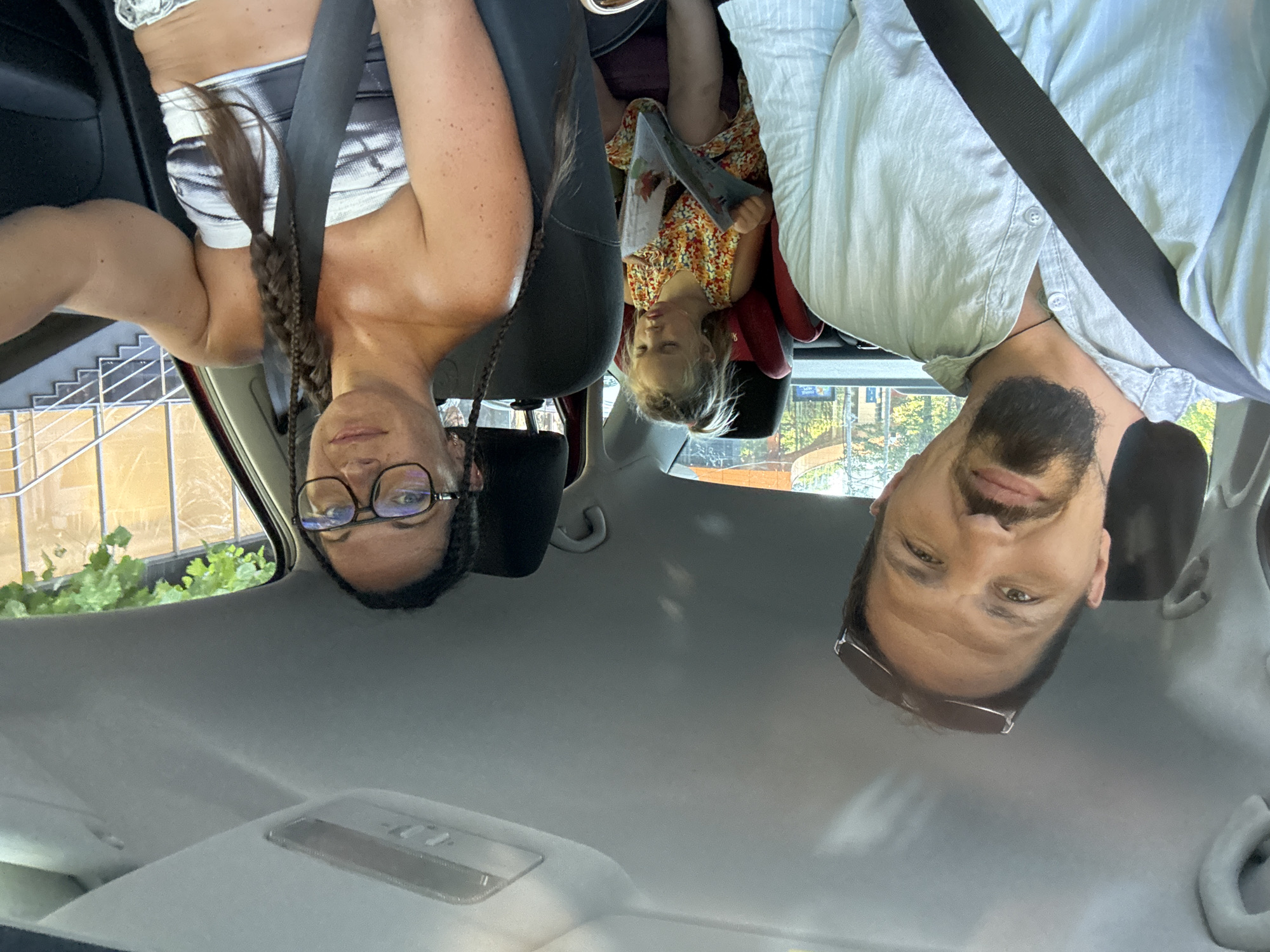 The image shows a family in a car, likely on a road trip. The photo is taken from the perspective of someone in the front passenger seat, looking back. Three people are visible: a woman in the front passenger seat, a man in the driver's seat, and a young child in a car seat in the back. The woman is wearing a black and white patterned top and has her hair in a braid. She is wearing glasses and has a serious expression. The man in the driver's seat is wearing a light blue shirt and sunglasses. He is smiling and looking at the camera. The child in the back is wearing a colorful dress and holding a book. The car appears to be moving, as indicated by the blurred background of trees and buildings. The time of day appears to be daytime, with sunlight filtering through the car windows. The overall mood is one of a happy family outing. The location is Sochi, Russia. No visible text is present in the image.
