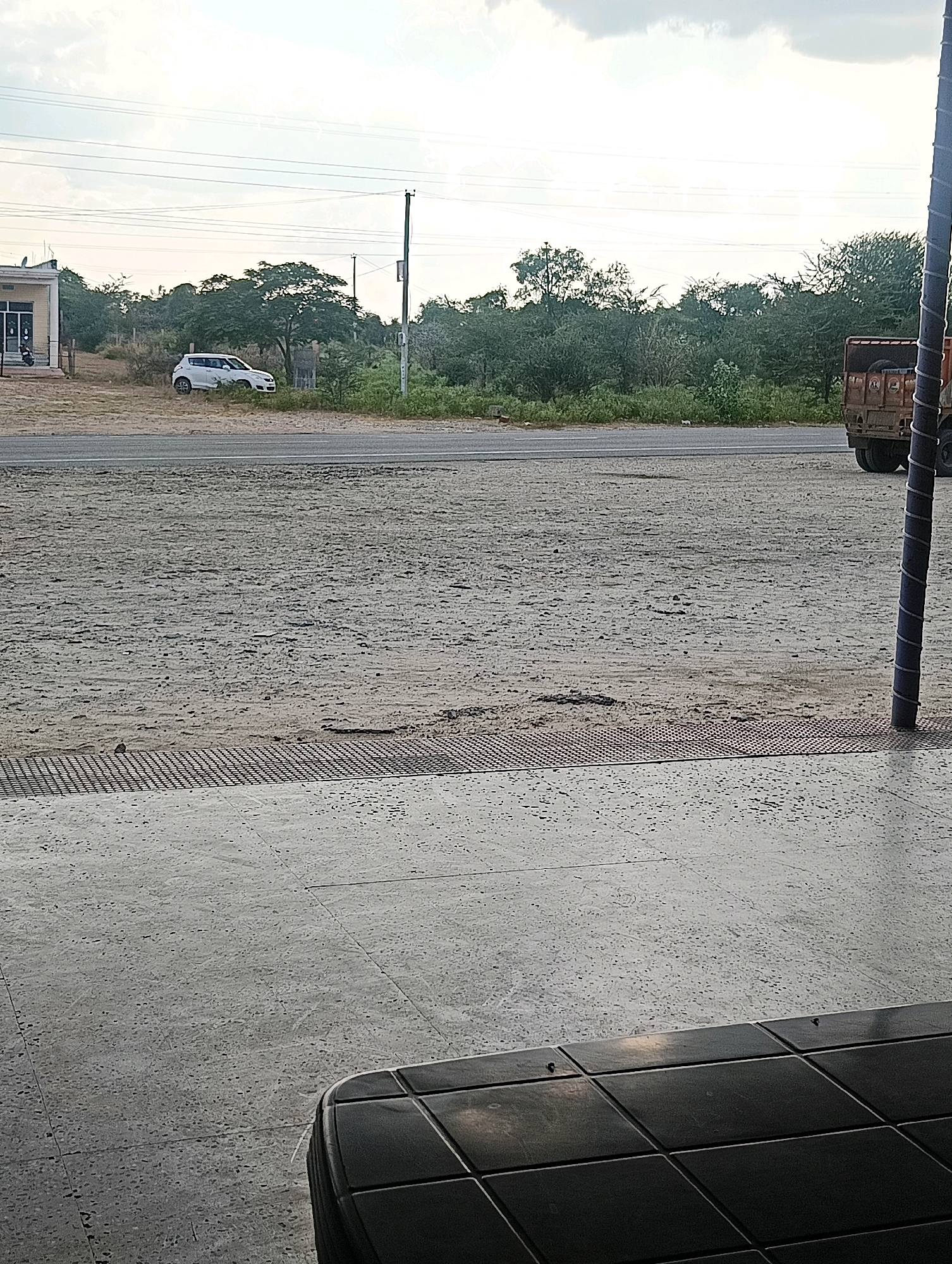 The image captures a scene in Chhapri Bari, India, focusing on a roadside area. In the foreground, a tiled surface with a dark, patterned area is visible. Beyond this, a paved surface with a metallic grate runs horizontally. Further back, a dusty, unpaved area leads to a paved road. On the road, a white car is parked on the right side, near a building. Across the road, a dense line of trees and vegetation forms the background, with a utility pole and wires stretching across the sky. To the far right of the image, the rear of a rustic brown truck is partially visible, parked on the side of the road. The sky is overcast with scattered clouds.