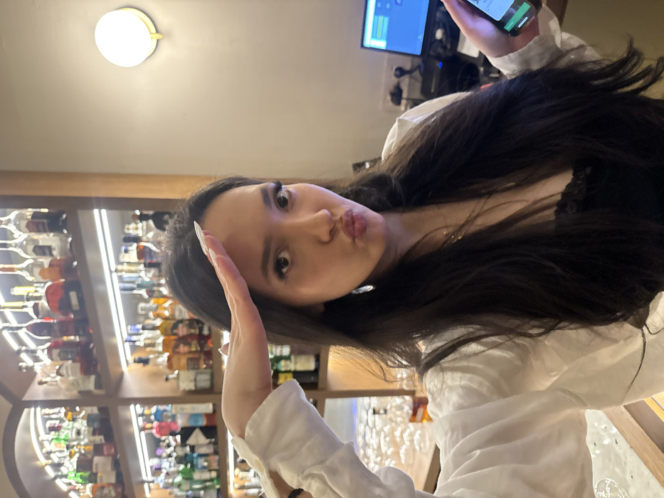 The image depicts a young adult female posing in a commercial establishment, specifically a bar or restaurant, located in Yerevan, Armenia. The subject, centered in the frame, has long dark hair and is wearing a white long-sleeved shirt over a black lace-trimmed top. Her left hand is raised to her forehead in a salute gesture, fingers extended, displaying long, light-colored fingernails. Her lips are pursed in a "duck face" expression. She wears a gold-colored necklace and a dangling earring on her visible left ear. In her right hand, partially visible, she holds a smartphone with an illuminated screen. The background to the left features a well-stocked bar with multiple shelves of various liquor bottles, illuminated by strip lighting. Below these shelves, a counter holds neatly arranged clear glassware. To the right of the subject, a light-colored wall displays a circular wall sconce providing warm illumination. Further right, a laptop or monitor with a blue display and associated electronic equipment are visible on a surface.
