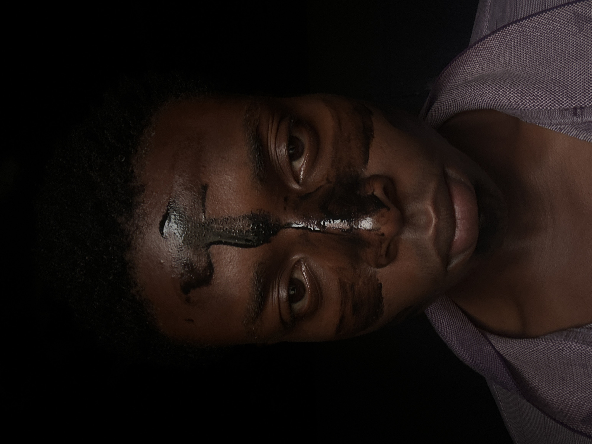 A close-up, low-light photograph shows a dark-skinned individual, likely male, with black streaks applied to their face. The streaks run across the forehead and down the nose. The person's eyes are open, and they appear to be wearing a shirt with a textured fabric. The background is black. There is no evidence to suggest this image was taken in Bukuru, Nigeria.