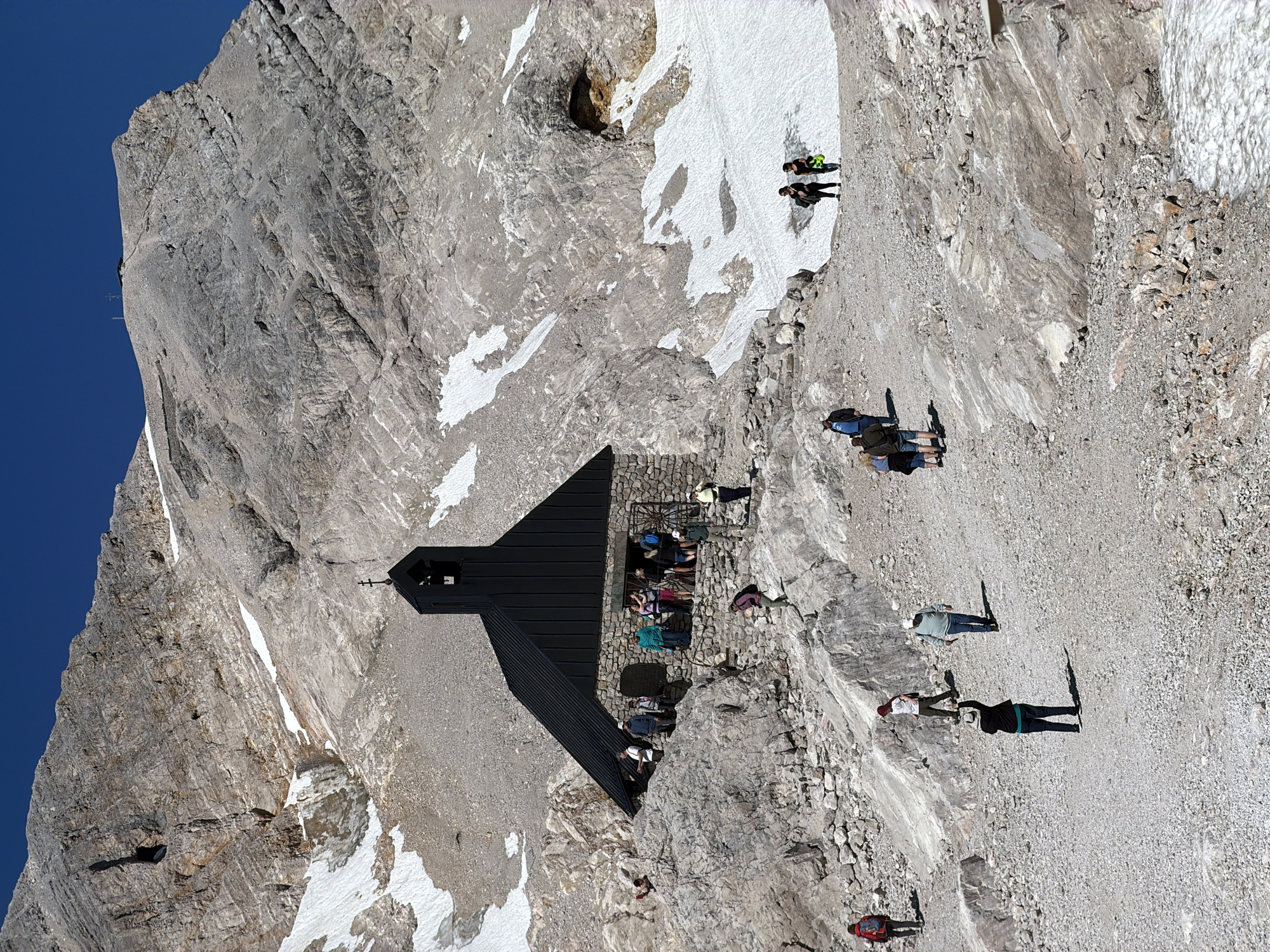 The image shows a remote mountain landscape with a small, dark-roofed chapel constructed of stone. The chapel is situated on a rocky, gravelly slope, with patches of snow visible in the background and on higher ground. Several people are scattered across the scene, some walking on the paths and others gathered near the chapel. The mountain terrain is rugged and appears to be at a high altitude, with a clear blue sky overhead. The location is the Zugspitze mountain, the highest peak in Germany, near Garmisch-Partenkirchen.