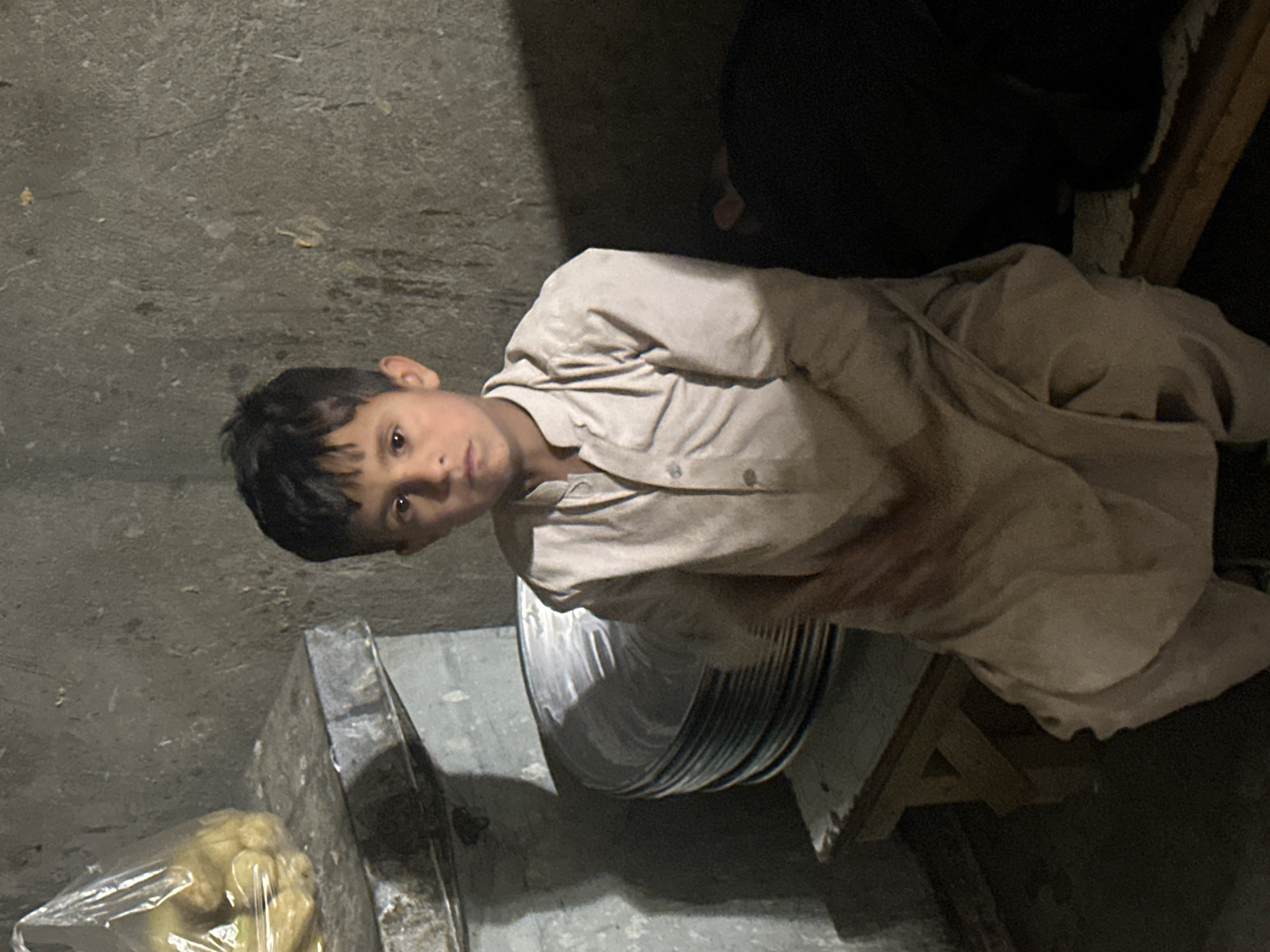 A young boy sits on a wooden stool in what appears to be an interior setting. The boy is wearing a light-colored, long-sleeved shirt. He is positioned next to a stack of metal plates. There is a plastic bag containing what looks like vegetables, possibly cauliflower, visible near a metallic surface. The background consists of a concrete wall.
The location is Quetta City Tehsil, Pakistan.