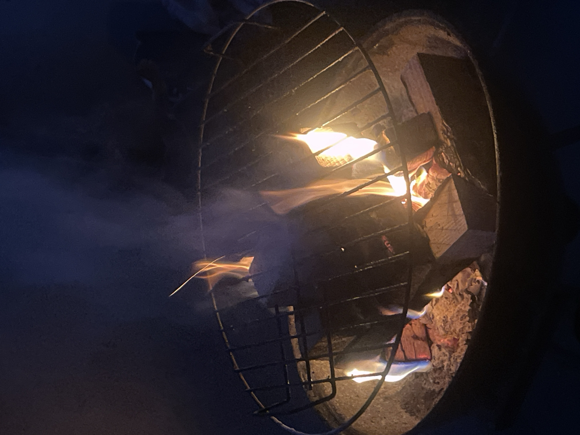 A close-up, low-angle shot captures a fire burning in a fire pit at night. A metal grate is suspended above the flames, which are licking at pieces of wood. Smoke rises from the fire, obscuring parts of the background. The fire pit appears to be made of concrete or stone, and the surrounding area is dark. The image is composed primarily of oranges, yellows, and blues, with the dark blue of the night sky dominating the upper portion. There are no people or animals visible in the frame.