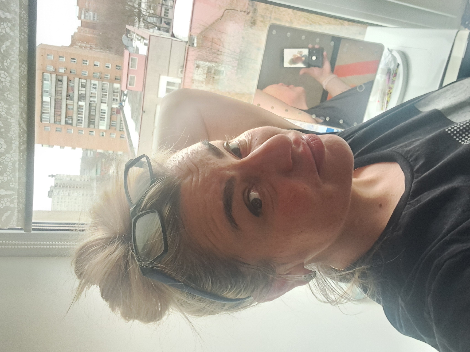 An adult female is captured in a close-up, eye-level photograph, positioned indoors near a window overlooking an urban landscape in Elektrostal, Russia. The subject has blonde or light-colored hair styled in a messy bun, with dark-framed reading glasses resting on top of her head. She wears a dark, possibly black, t-shirt with a subtle pattern of light dots on the lower left front. A small silver-colored earring is visible in her right ear. Her left arm is raised, with her hand positioned towards the back of her head. She is looking directly at the camera with a slight pout.
To her right, a window allows natural light into the scene. A translucent white, patterned roller blind with a beaded pull cord partially covers the top of the window. On the windowsill, a rectangular lighted makeup mirror reflects a partial image of the woman's right arm, adorned with a light-colored wristband, holding a black smartphone, indicating she is taking the photograph. Small, elongated objects, possibly pens or brushes, are also present on the sill. The interior wall to her left is plain white.
Through the window, the outdoor environment of Elektrostal, Russia, is visible. A prominent multi-story residential building, constructed of orange-brown brick or concrete, with numerous windows and balconies, dominates the background. Below this building, a flat rooftop of a smaller structure is visible, partially covered with snow, featuring a small pink rectangular enclosure with two windows. Bare deciduous trees are sparsely distributed throughout the urban landscape, suggesting a winter or early spring season under an overcast sky.