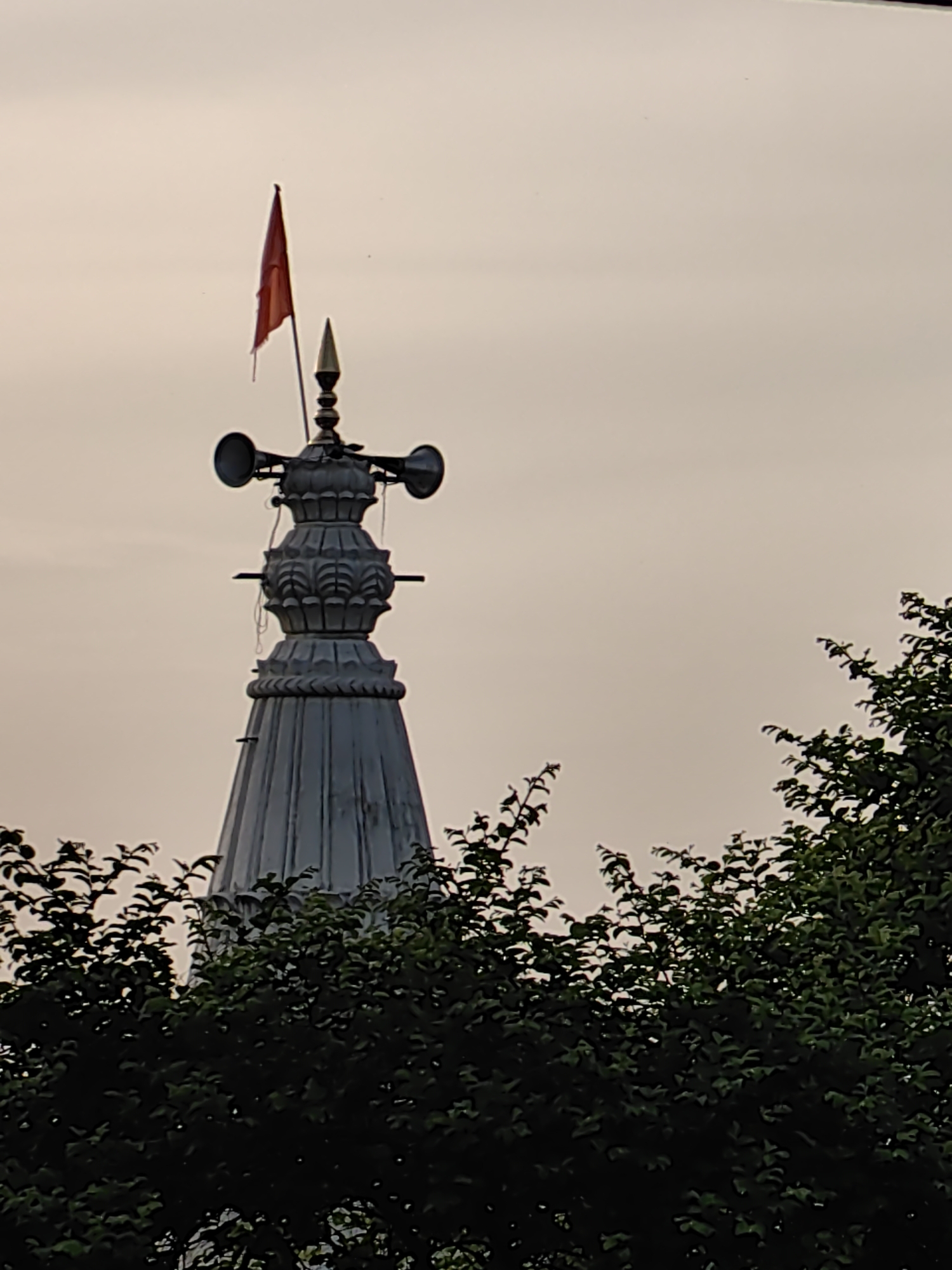 The image captures the upper portion of a religious structure, likely a temple spire, in Mhaiswadi, India. A prominent red flag flutters from a flagpole atop the spire. Two loudspeakers are mounted on the structure, one on each side, at the middle section. The spire itself is ornately decorated with multiple tiers and intricate carvings. Below the spire, dense green foliage from trees obscures the lower part of the structure and extends across the foreground, suggesting the building is surrounded by vegetation. The sky is overcast with a pale, muted tone, indicating either dawn, dusk, or a heavily cloudy day. There are no people visible in the frame.