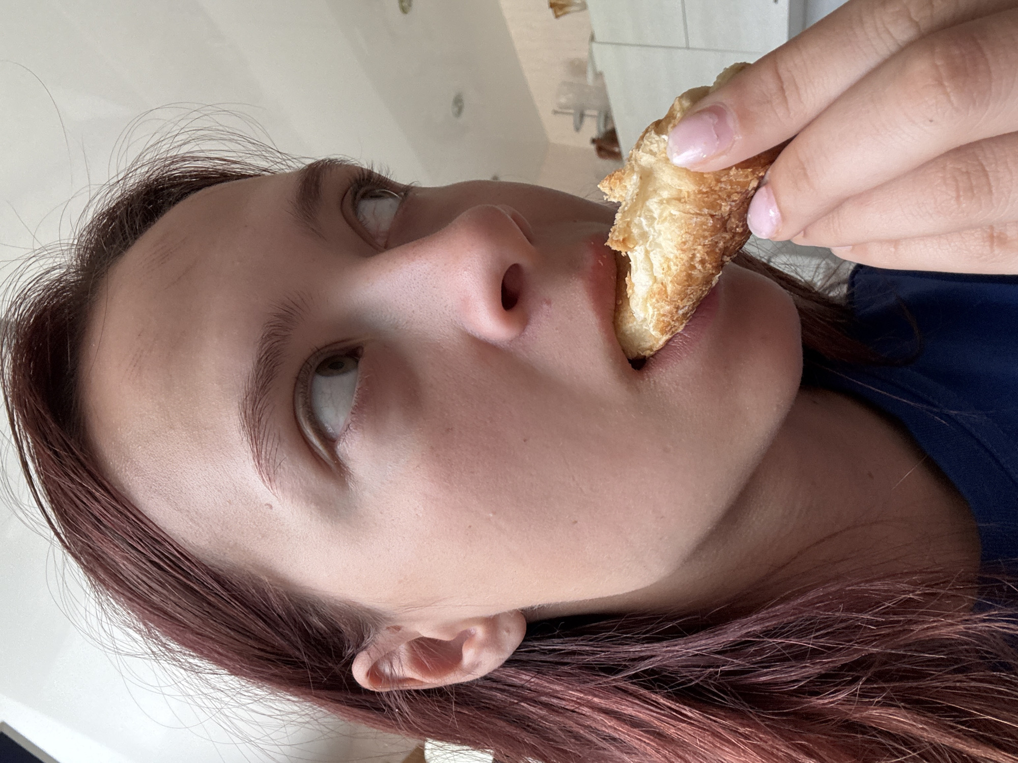 The image presents a close-up, upward-angled view of a person in the act of eating. The subject, who appears to be female, has fair skin and reddish-brown hair. Her eyes are cast upwards and to the right, suggesting she is either focused on the pastry or looking at something above.
She is holding a golden-brown, flaky pastry, resembling a croissant or similar baked good, which is partially in her mouth. Her hand, with natural-looking fingernails adorned with a light pink polish, firmly grips the pastry. A portion of a dark blue garment, likely a shirt or top, is visible on her right shoulder.
The background is out of focus and indistinct, showing light-colored walls and ambiguous shapes that suggest an indoor environment, such as a home or café. The lighting is soft and ambient, providing no specific clues about the time of day. There is no visible text in the image. The scene captures a mundane, personal moment of enjoying food, possibly in a domestic setting within Прикамский, Russia, though no specific geographical identifiers are visible in the photograph itself.