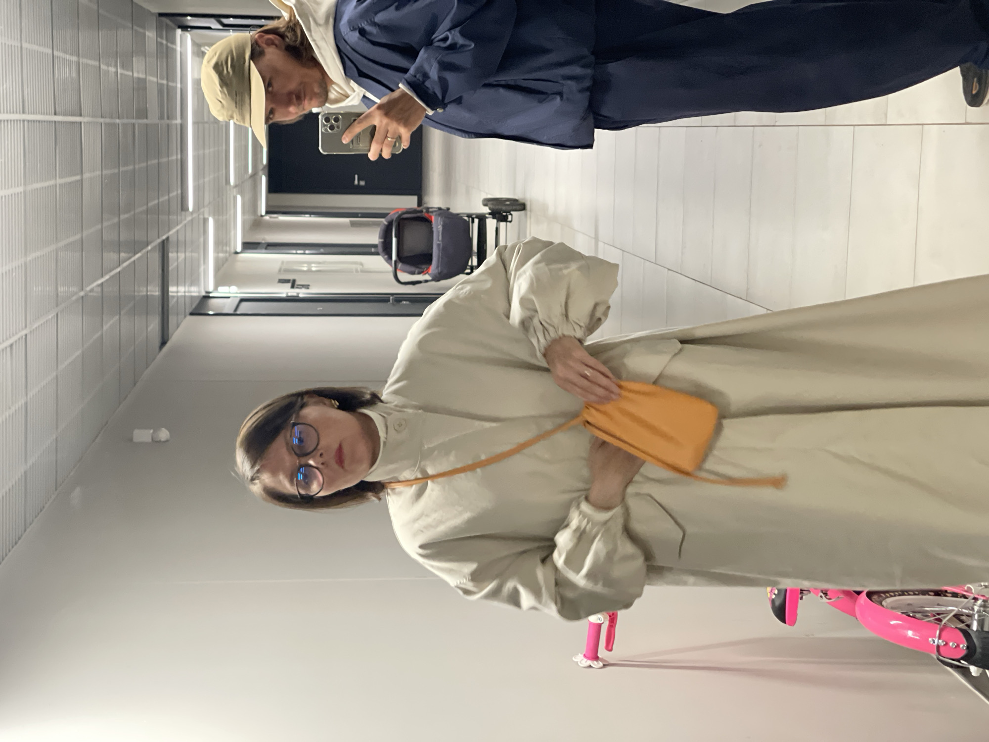 Two individuals stand in a well-lit corridor. One, wearing a light beige trench coat and glasses, holds an orange bag. A child's pink bicycle is partially visible in the bottom right. The other individual, wearing a navy blue jacket and beige hat, is holding a mobile phone and appears to be taking a picture. A stroller is visible in the background. The corridor has white walls and a grid-like ceiling. The location is Unknown, Unknown.