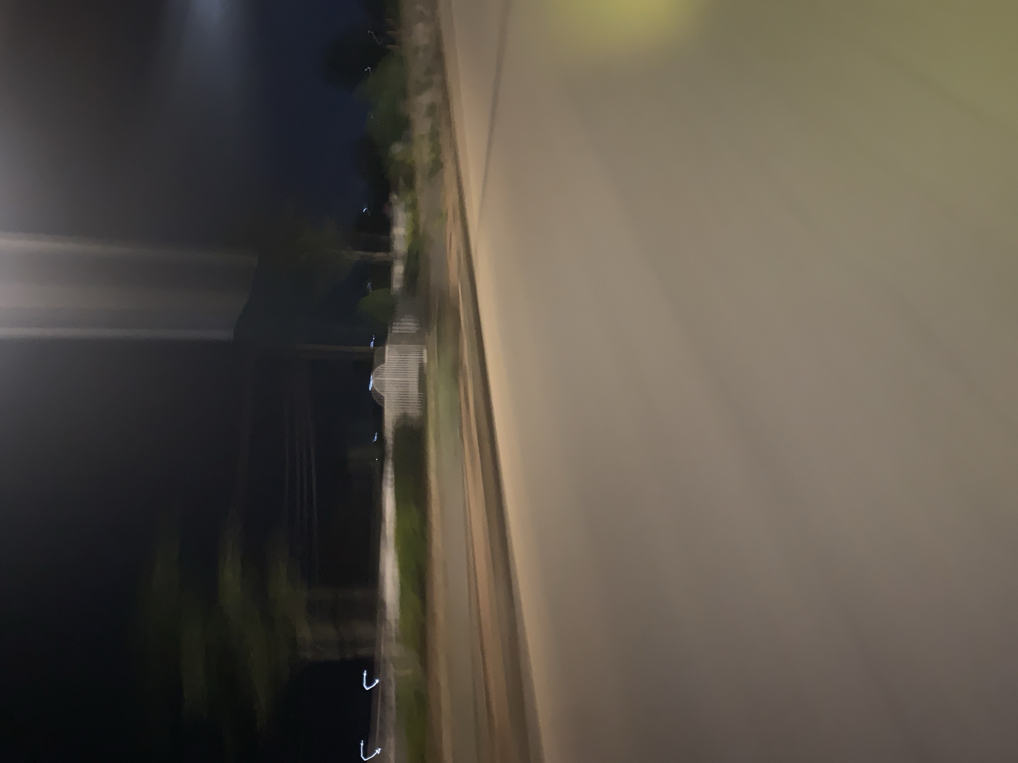The image depicts a blurred, nighttime street scene in Bukuru, Nigeria. The foreground is dominated by a paved road, appearing light brown and smooth. To the left, a white fence is visible, suggesting a boundary or property line. Beyond the fence, faint green foliage and trees can be discerned, interspersed with a dark, indistinct background that signifies the night sky. Several small, bright blue lights, possibly streetlights or vehicle lights, are scattered along the distant horizon, indicating the presence of a populated area or roadway. The overall impression is one of motion, likely due to the camera's movement, creating streaks of light and blurring the details of the environment. There are no discernible people or specific actions taking place.