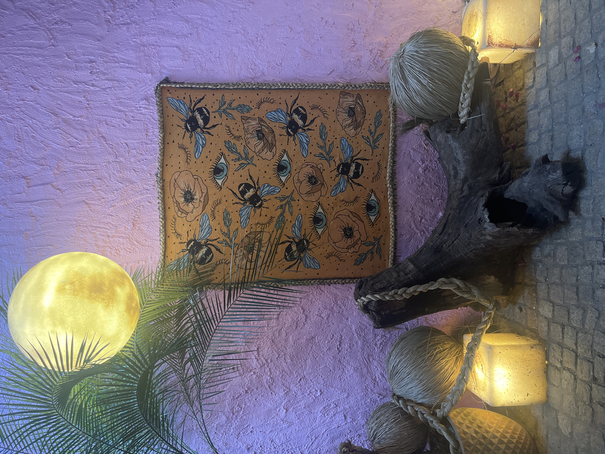 A horizontally oriented, medium shot shows an interior wall with decorations. The wall has a heavily textured surface with a lavender and pink color. A square textile is hanging on the wall, with a border of woven material. The textile features a pattern of bees, flowers, and eyes in shades of brown and blue. To the right of the textile is a piece of driftwood with twine wrapped around it, and two straw balls sitting on top. A string of cube-shaped lights is wrapped around the driftwood, and extends to the right side of the image. To the left of the textile is a leafy plant and a spherical light. The background of the image is a lavender and pink textured wall. The image appears to be taken indoors. There is no indication of the city or location. No people are visible in the image.