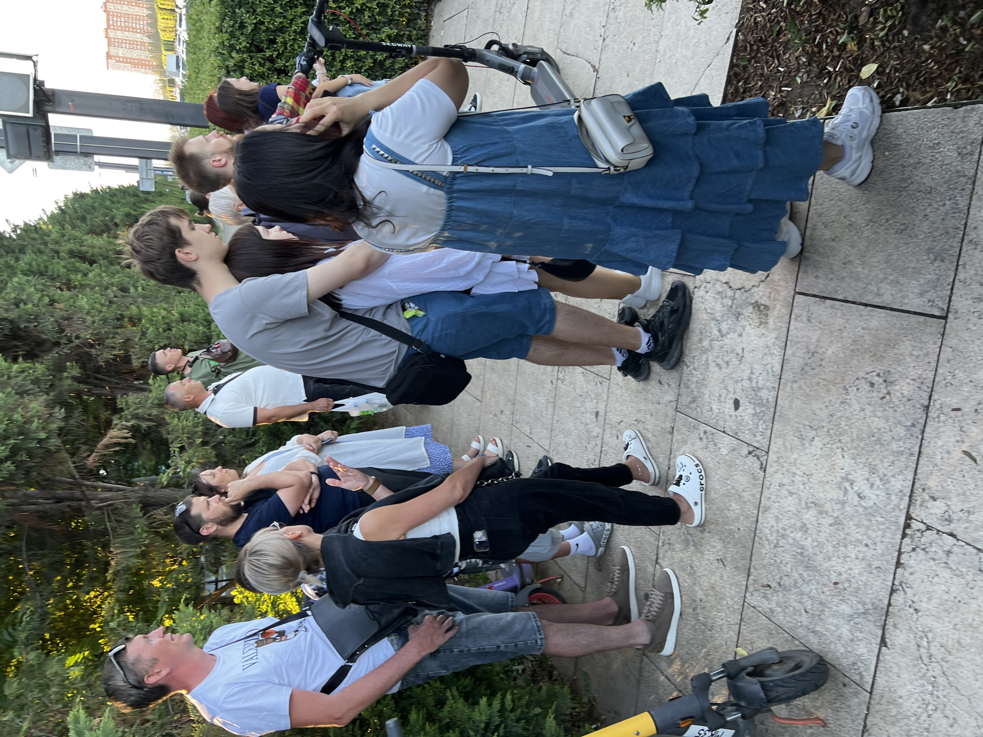 The image depicts a group of people standing on a paved walkway lined with trees and bushes, with a street and buildings visible in the background. The overall setting suggests an outdoor urban environment. Several individuals are facing away from the camera, some interacting with each other. One person on the left is wearing a white t-shirt and grey shorts, and has a bag slung over their shoulder. A woman next to them is wearing black pants and a black jacket draped over her shoulders, and white clogs. Another woman in a white dress is also visible. Further down the walkway, a group of younger people are standing together. One person in a grey t-shirt is with a person in a white shirt and blue shorts, and another individual in a blue denim dress is holding a white purse. Two electric scooters are present on the walkway, one in the foreground on the left and another further back on the right. The background shows a street with parked cars and a tall building with multiple floors. The lighting suggests it is daytime, likely late afternoon or early evening, given the shadows. The environment appears to be a public space in the city of Krasnodar, Russia, potentially a park, promenade, or pedestrian area.
