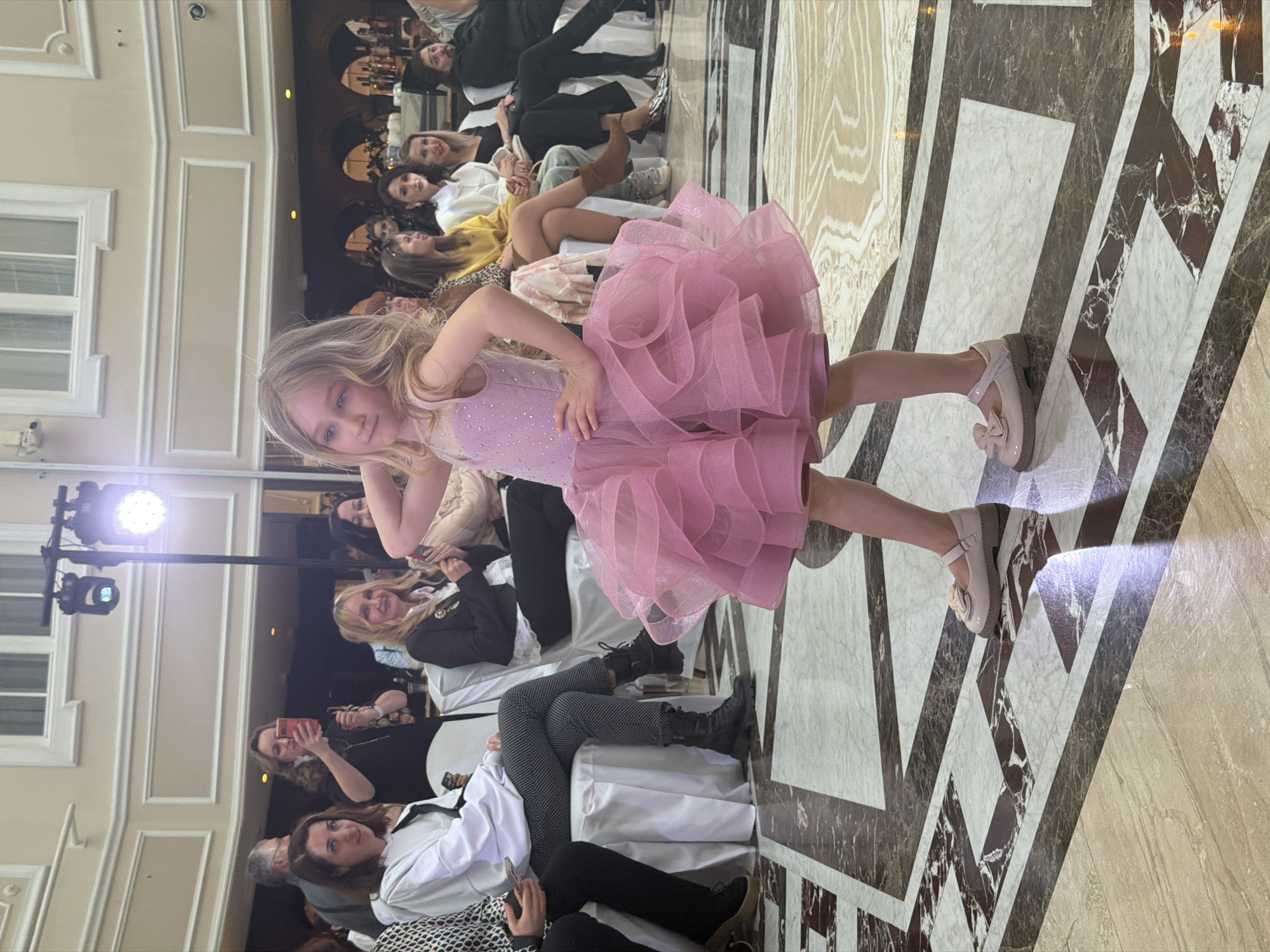 A young girl in a pink dress poses for a photograph on a patterned floor, likely inside a building due to the interior architecture visible. Several people are seated in the background, seemingly watching or attending the event. A spotlight illuminates the scene. The location is not discernible as Saint Petersburg, Russia based on visual information.