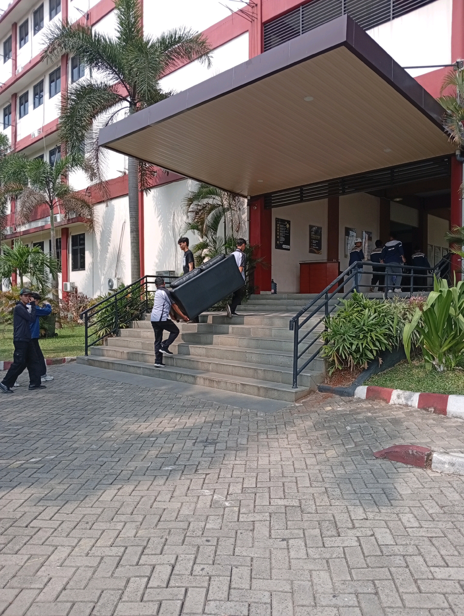 In Kelapa Dua, Indonesia, a group of individuals are seen carrying a large black object, possibly a sofa or a piece of furniture, up a set of outdoor stairs leading to a building entrance. The building appears to be a multi-story structure with white walls and red trim. Palm trees and other greenery are visible around the building, suggesting a tropical or subtropical climate. The ground in front of the stairs is paved with interlocking bricks. One person in the foreground is holding a camera, appearing to take a photo or video. The scene depicts a logistical task of moving an object into or out of the building.