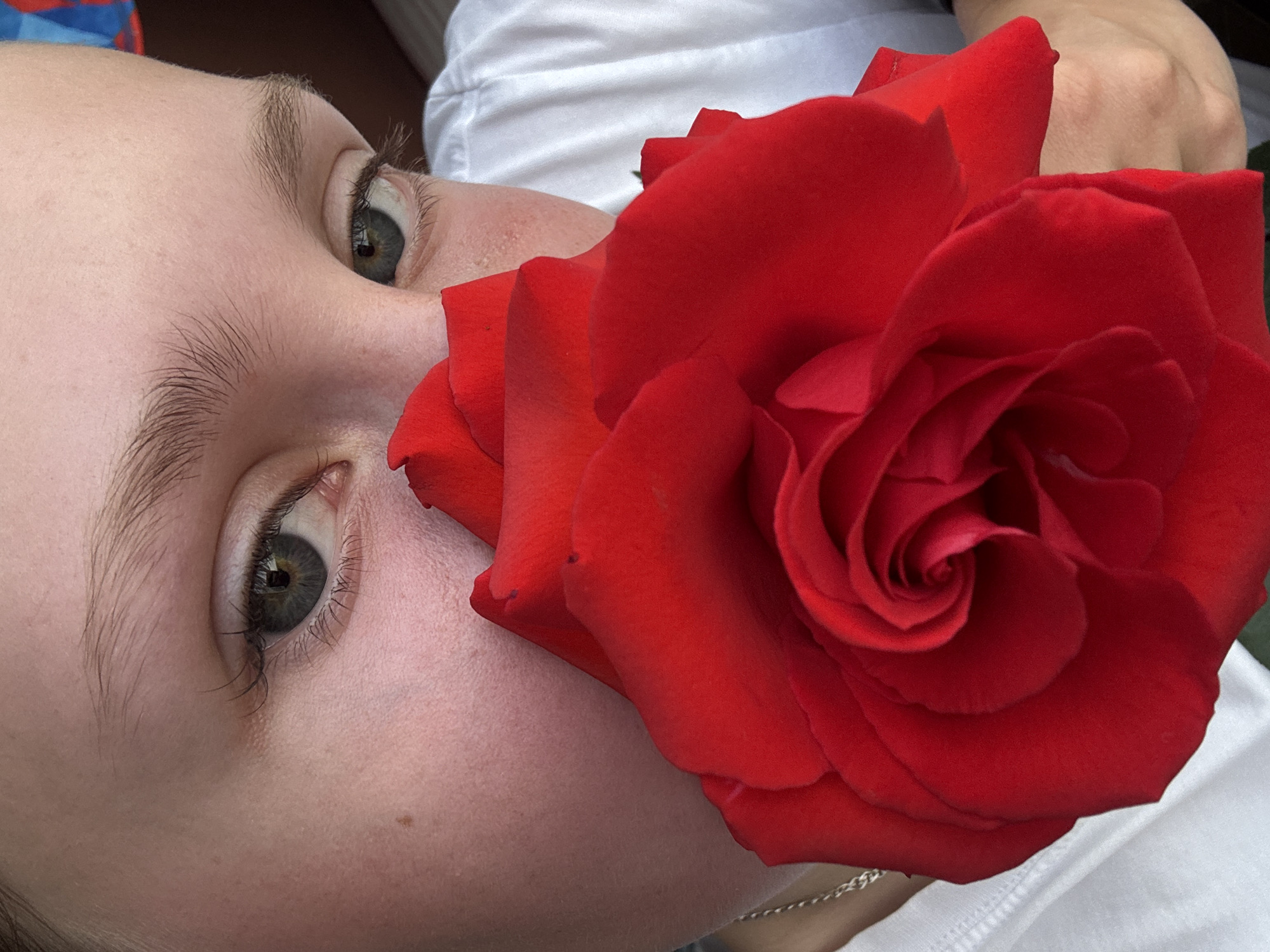 This close-up portrait captures the left side of a person's face closely interacting with a large, vibrant red rose. The subject has fair skin, with finely detailed eyebrows and long, delicate eyelashes framing a captivating grey-green eye that gazes upwards. A small brown mole is visible on the cheek below the eye.
A magnificent, fully bloomed red rose dominates the right side of the frame, its numerous velvety petals intricately folded. The rose appears to be held or placed intimately close to the person's face, touching their cheek and extending towards the nose, creating a striking contrast between the soft skin and the rich crimson of the petals. The lighting is bright and natural, suggesting a daytime setting.
The background is softly blurred, revealing hints of white fabric, likely a shirt, and other indistinct, darker elements, ensuring the focus remains entirely on the face and the flower. No specific location cues are visible within the image itself, beyond the provided context of Chaikovsky, Russia. There is no visible text. The overall impression is one of serene beauty and intimacy, highlighting the delicate features of the person and the lushness of the rose.