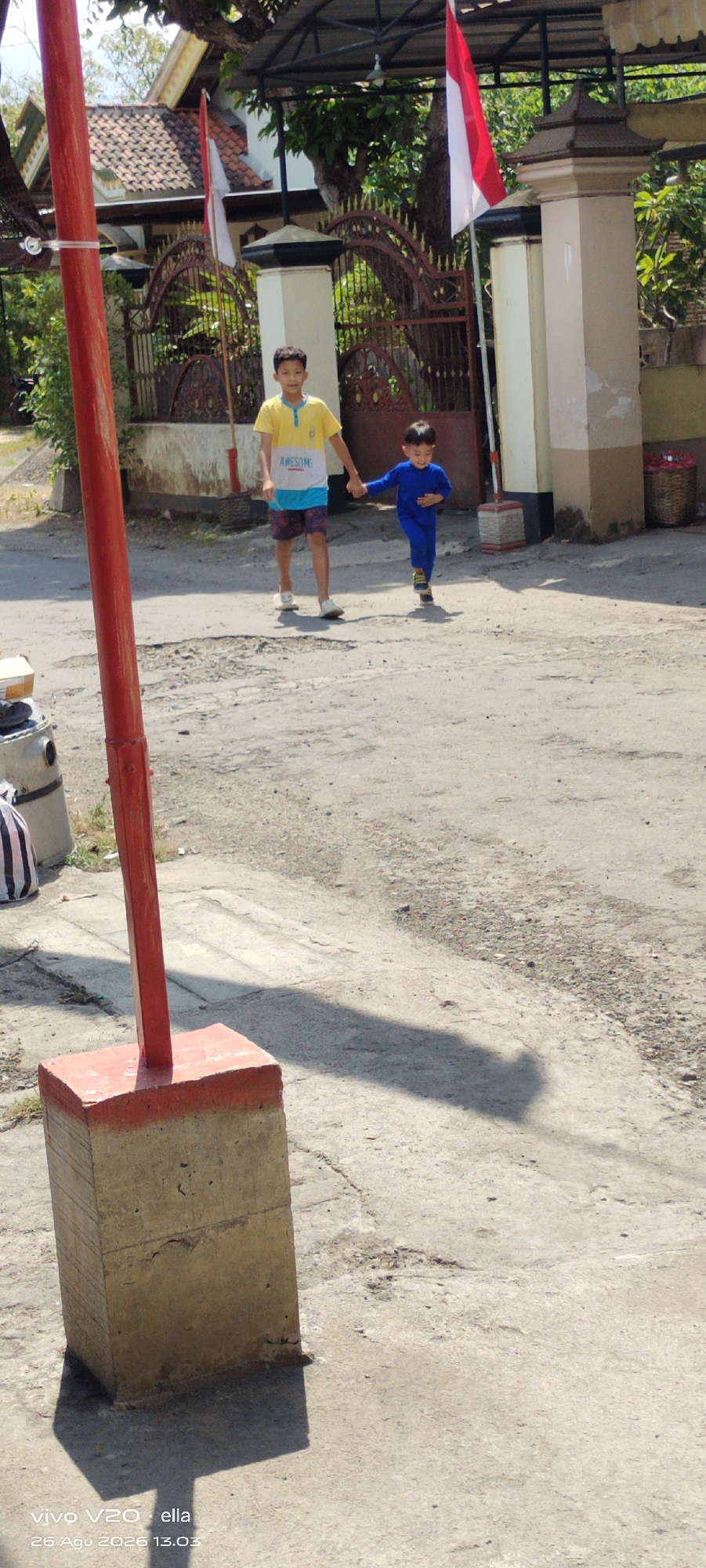 In Simo, Indonesia, two young boys walk hand-in-hand on a paved outdoor area. The boy on the left, wearing a yellow and blue t-shirt with the word "AWESOME" printed on it, and patterned shorts, is being led by the boy on the right, who is dressed in a full blue jumpsuit. They are walking towards the right side of the frame.
In the background, a house with a red-tiled roof is visible, adorned with Indonesian flags. A decorative metal gate separates the house from the street. A tall, red pole stands prominently in the foreground on the left, casting a long shadow across the ground. The ground itself appears to be a mix of concrete and asphalt, with some cracks and imperfections. Sunlight illuminates the scene, creating distinct shadows. A partially visible gray cylindrical object with black stripes is also present in the lower left corner. The overall environment suggests a sunny day in a residential area.
